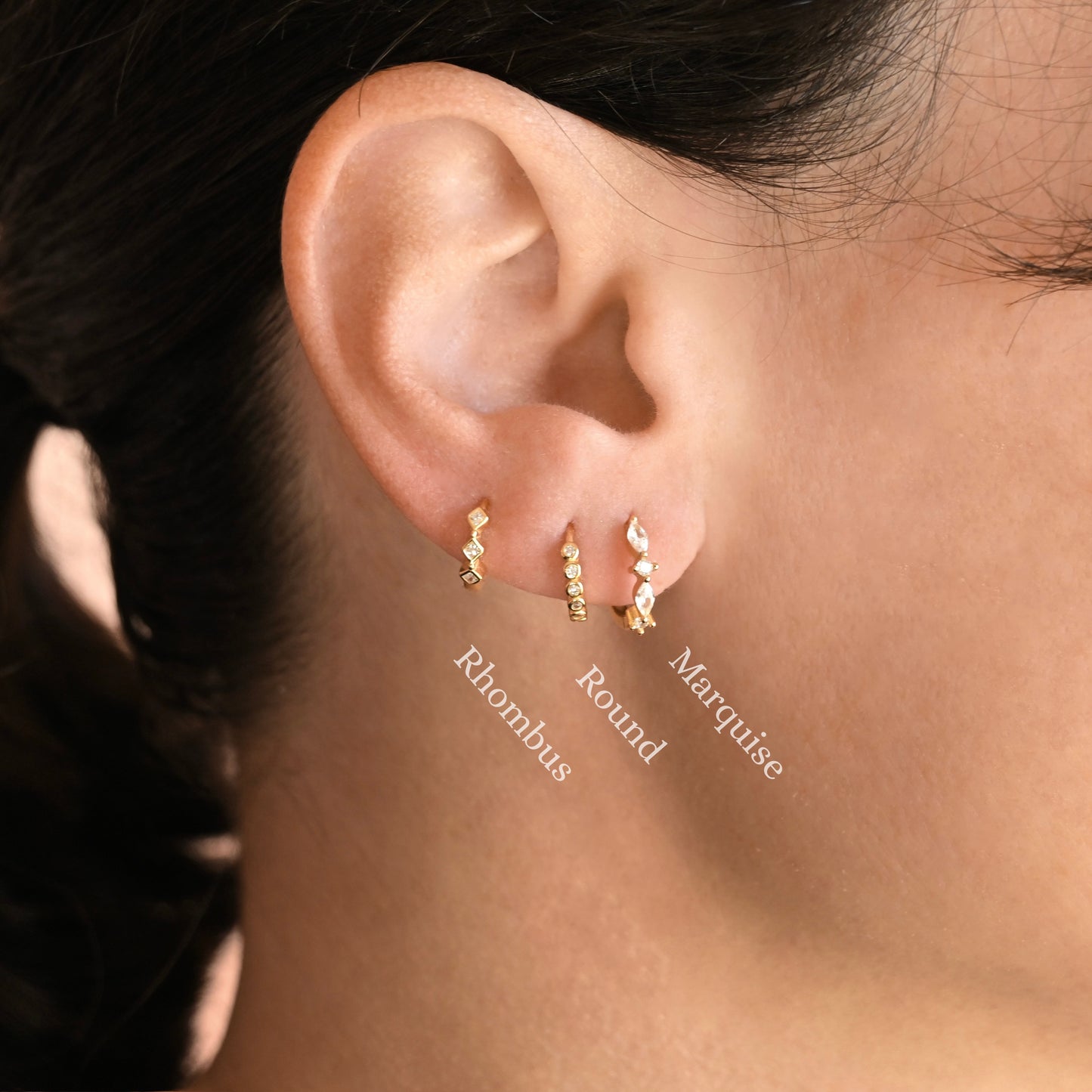 Close-up of a person's ear wearing three small gold ear cuffs labeled Rhombus, Round, and Marquise, with the names overlaid on the photo.