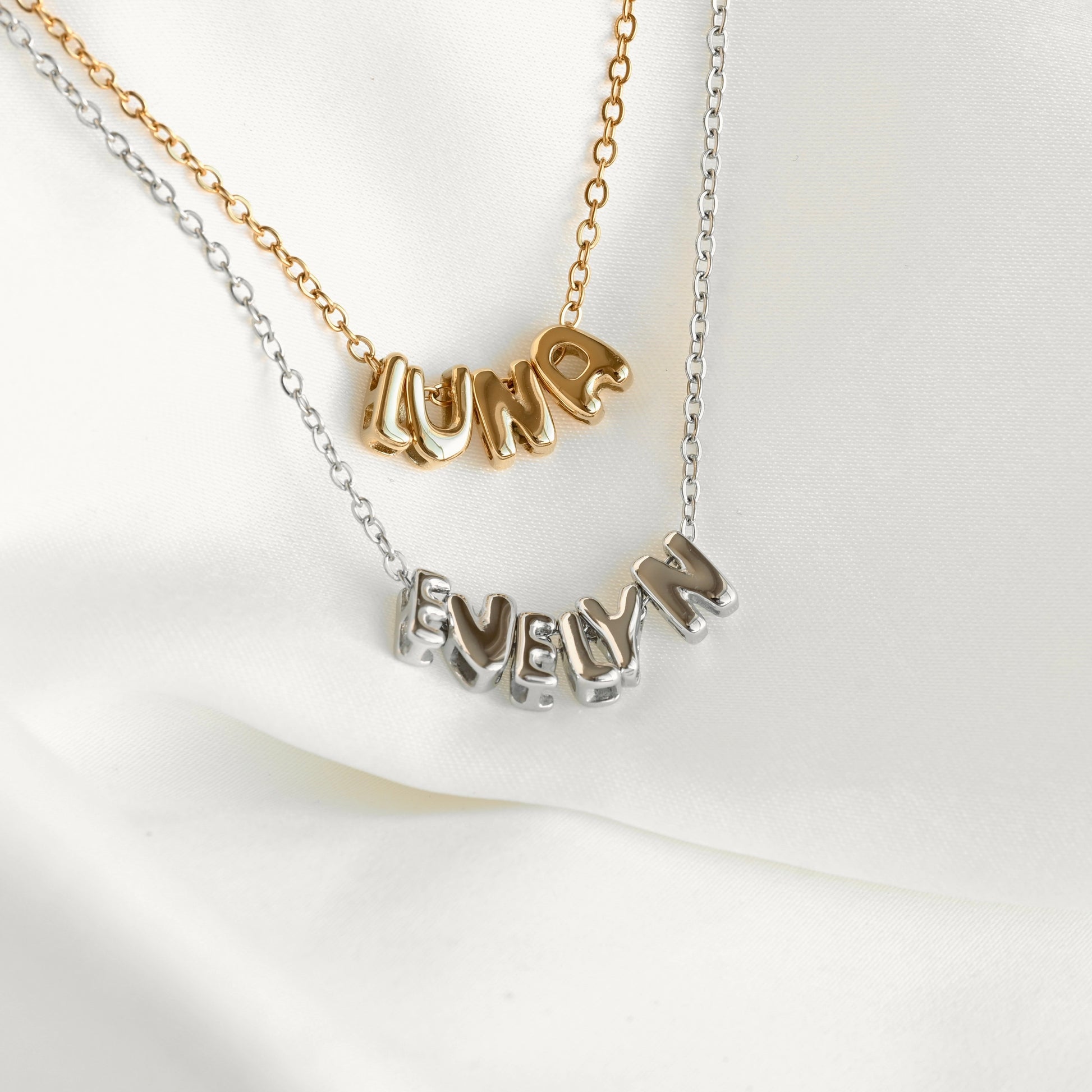 Two personalized name necklaces displayed on a soft white fabric background. One necklace has a gold chain with a gold nameplate spelling 'LUNA' and one in silver spalling 'EVELYN'.