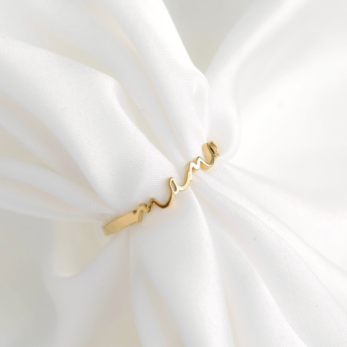 A delicate gold ring with a wavy script design spelling 'mama', displayed on soft, draped white fabric.