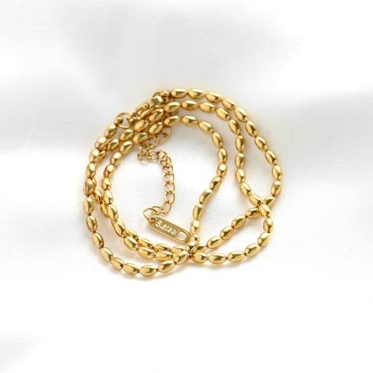 A gold chain necklace with elongated oval link design, coiled on a white fabric background. A small rectangular tag reading 'S.steel' is attached to the chain.
