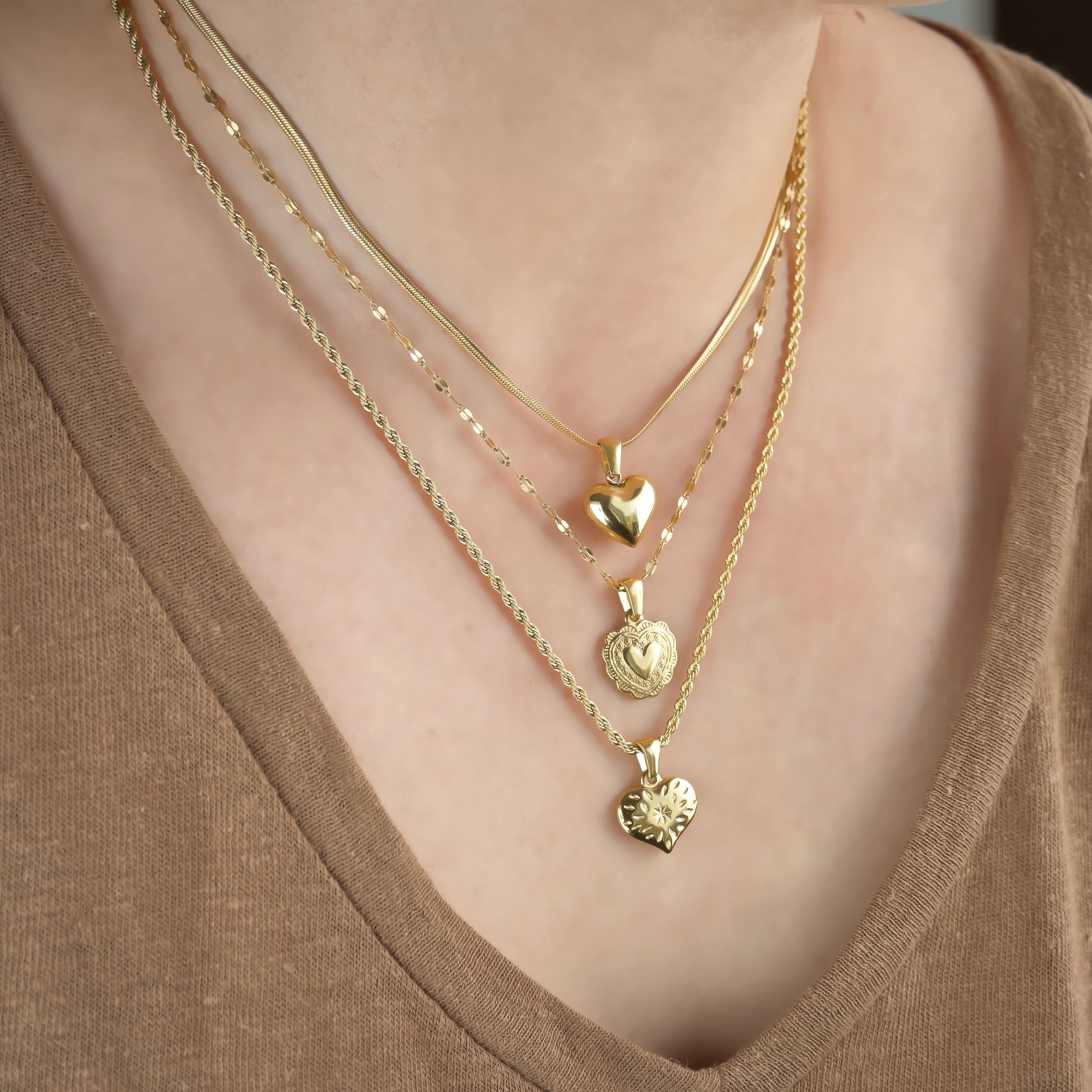 Close-up of a person wearing multiple gold necklaces with heart pendants: a flat snake chain with puffed heart, a dainty mariner chain with scalloped heart, and a rope chain with a star burst engraved heart.