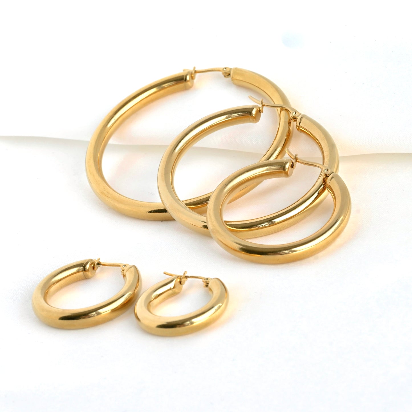 Gold hoop earrings on a white background, with two small hoops in the front and 3 larger sized hoops on the back standing against a folded satin fabric