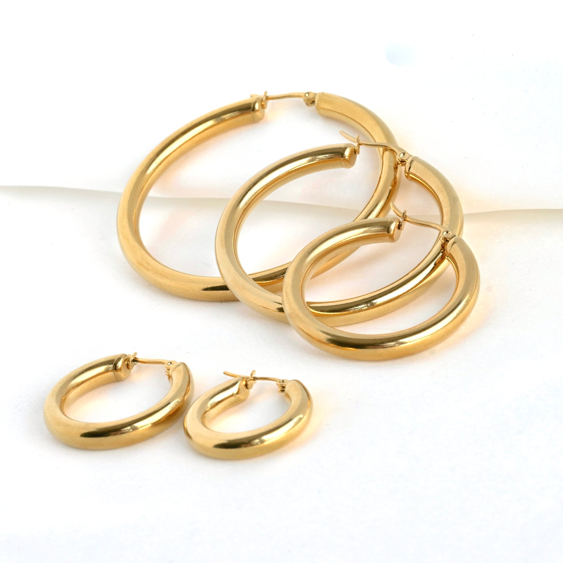 Gold hoop earrings on a white background, with two small hoops in the front and 3 larger sized hoops on the back standing against a folded satin fabric