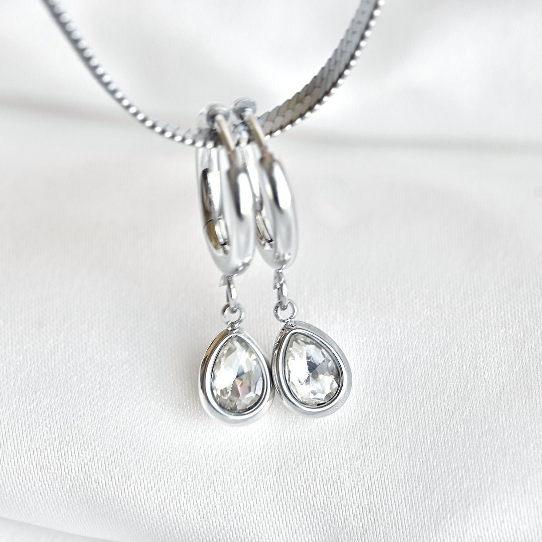 A pair of silver hoop earrings with dangling teardrop-shaped clear gemstones, hanging from a silver chain against a white fabric background.