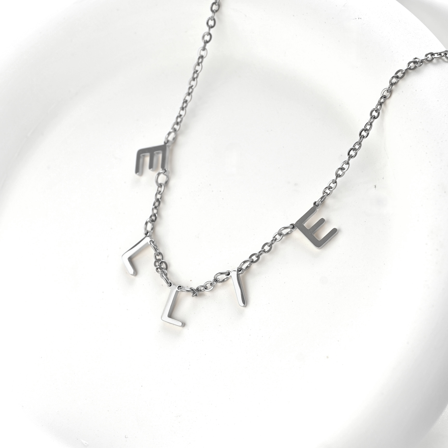 A silver initial necklace spelling 'ELLIE' on a delicate chain, displayed on a white ceramic dish.