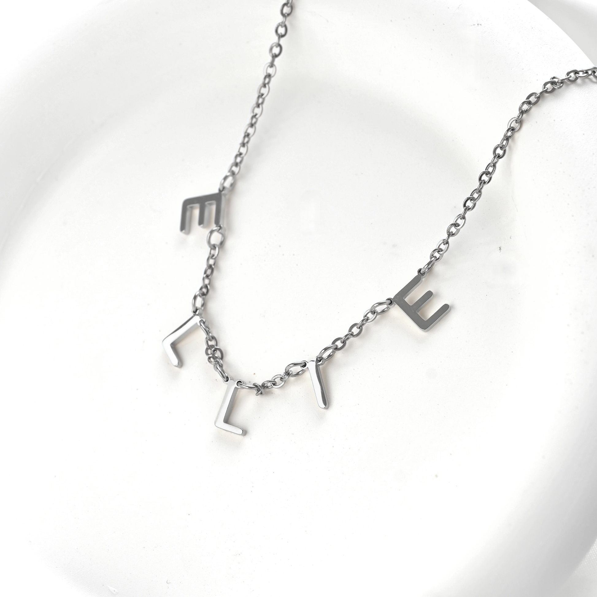 A silver initial necklace spelling 'ELLIE' on a delicate chain, displayed on a white ceramic dish.