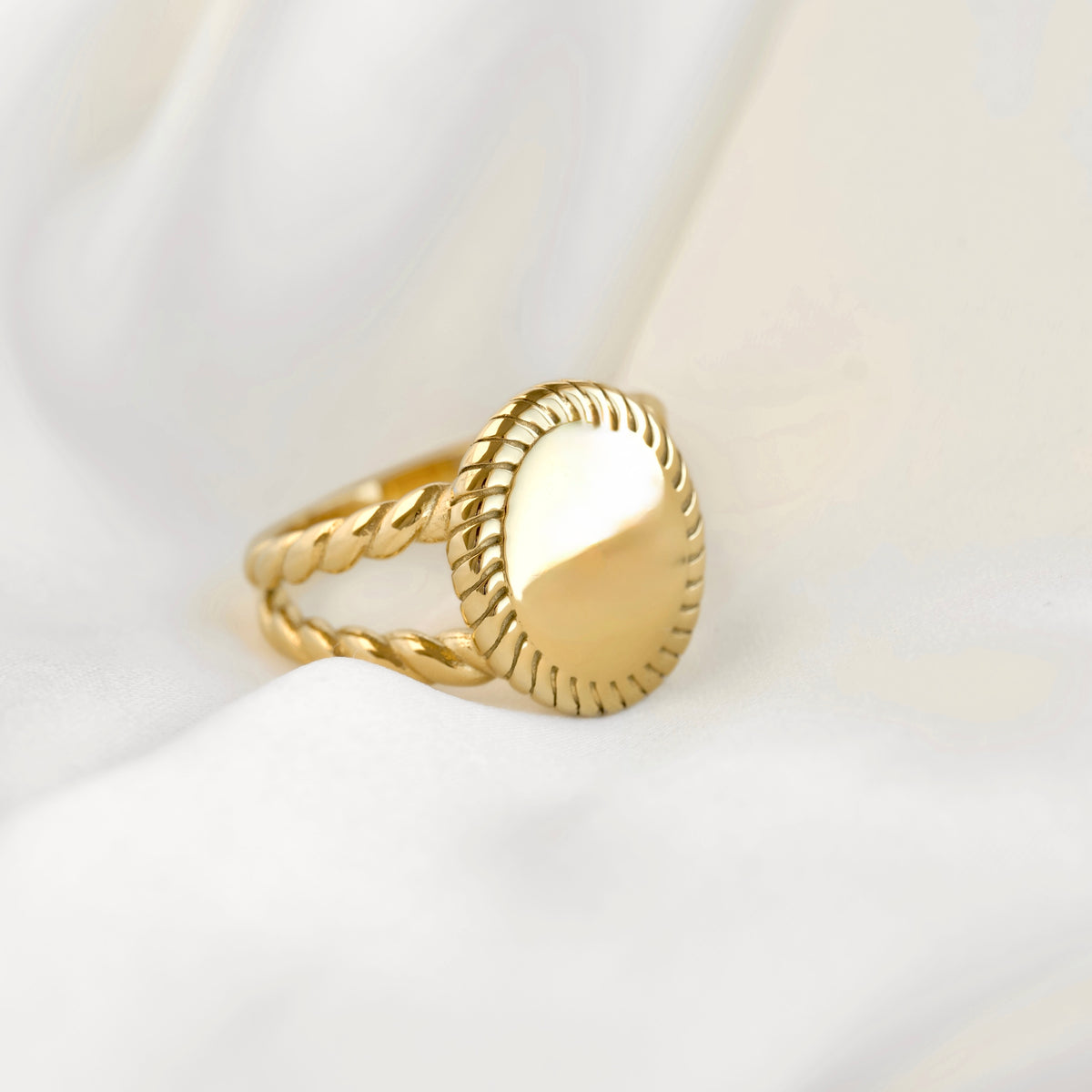 A gold ring with a twisted rope-style band and a smooth oval face, resting on draped white fabric.
