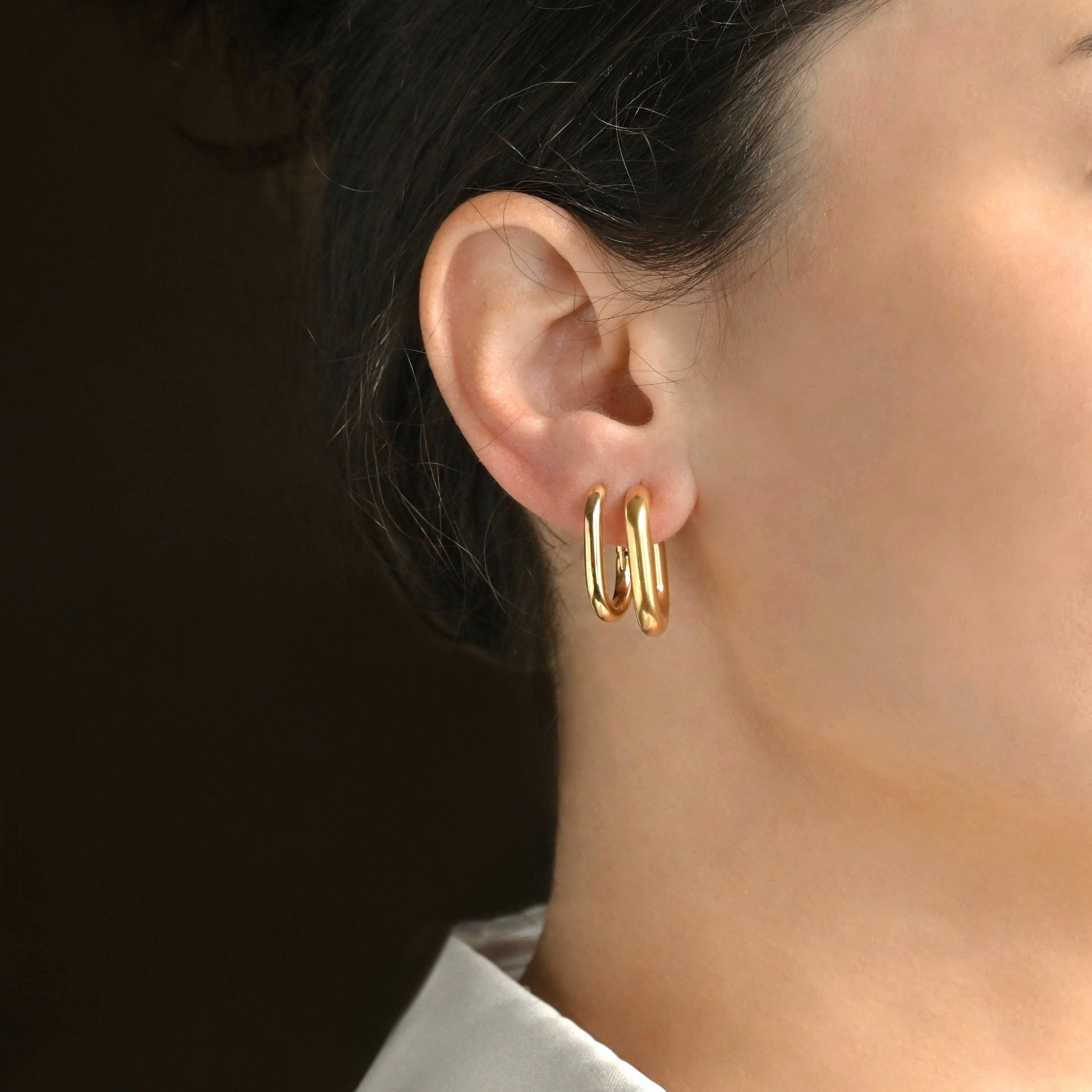 Pair of medium size gold U-shape earrings, one thick and one dainty worn by a person with dark hair.
