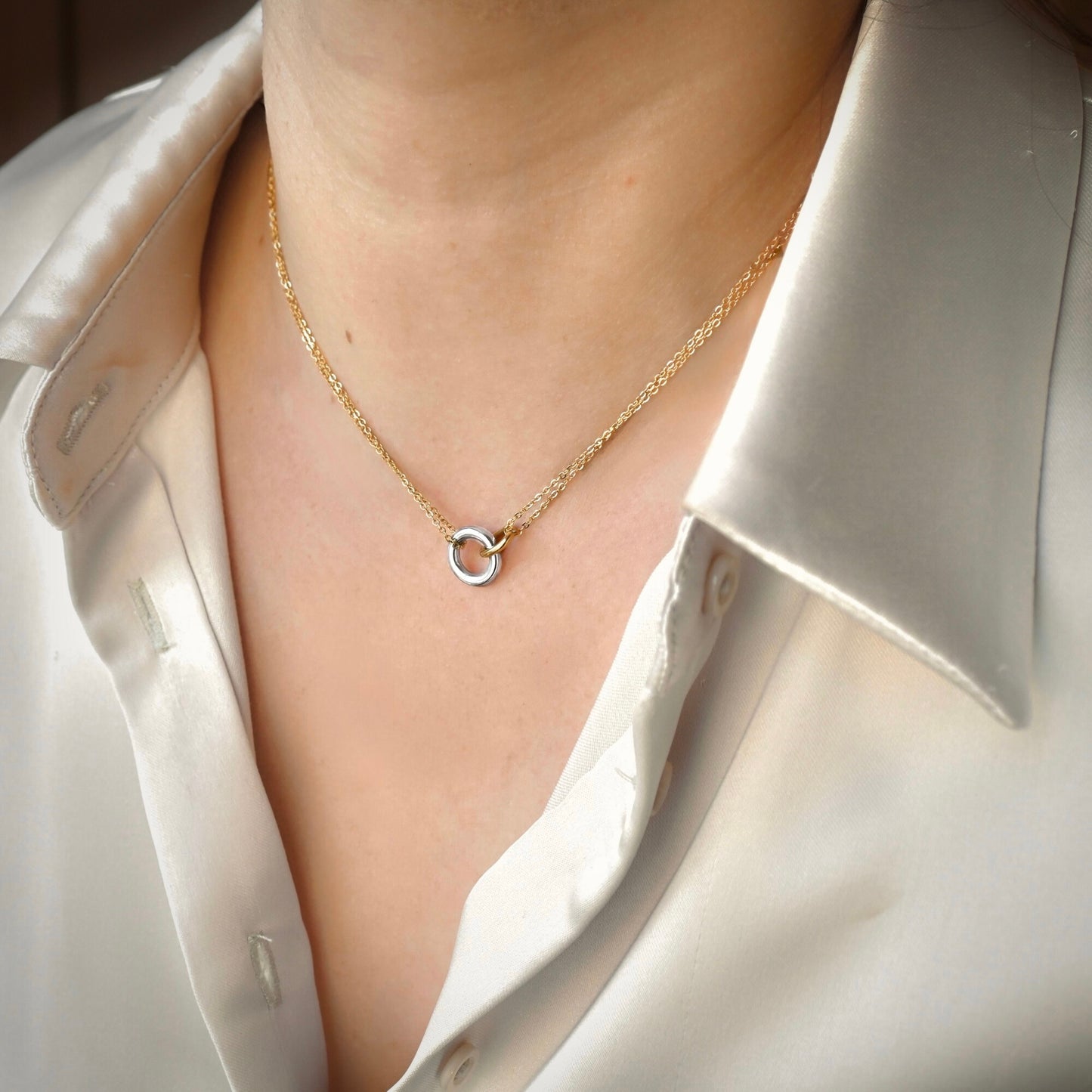 A close-up of a person wearing a delicate gold chain necklace with one medium silver and one oval gold interlocking rings, styled with a white satin collared shirt.
