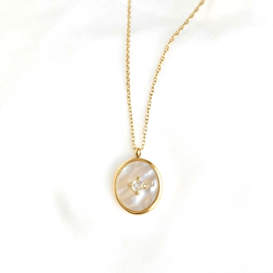 A gold chain necklace with an oval pendant featuring a white mother-of-pearl inlay and a small clear gemstone at the center, displayed on a white fabric background.