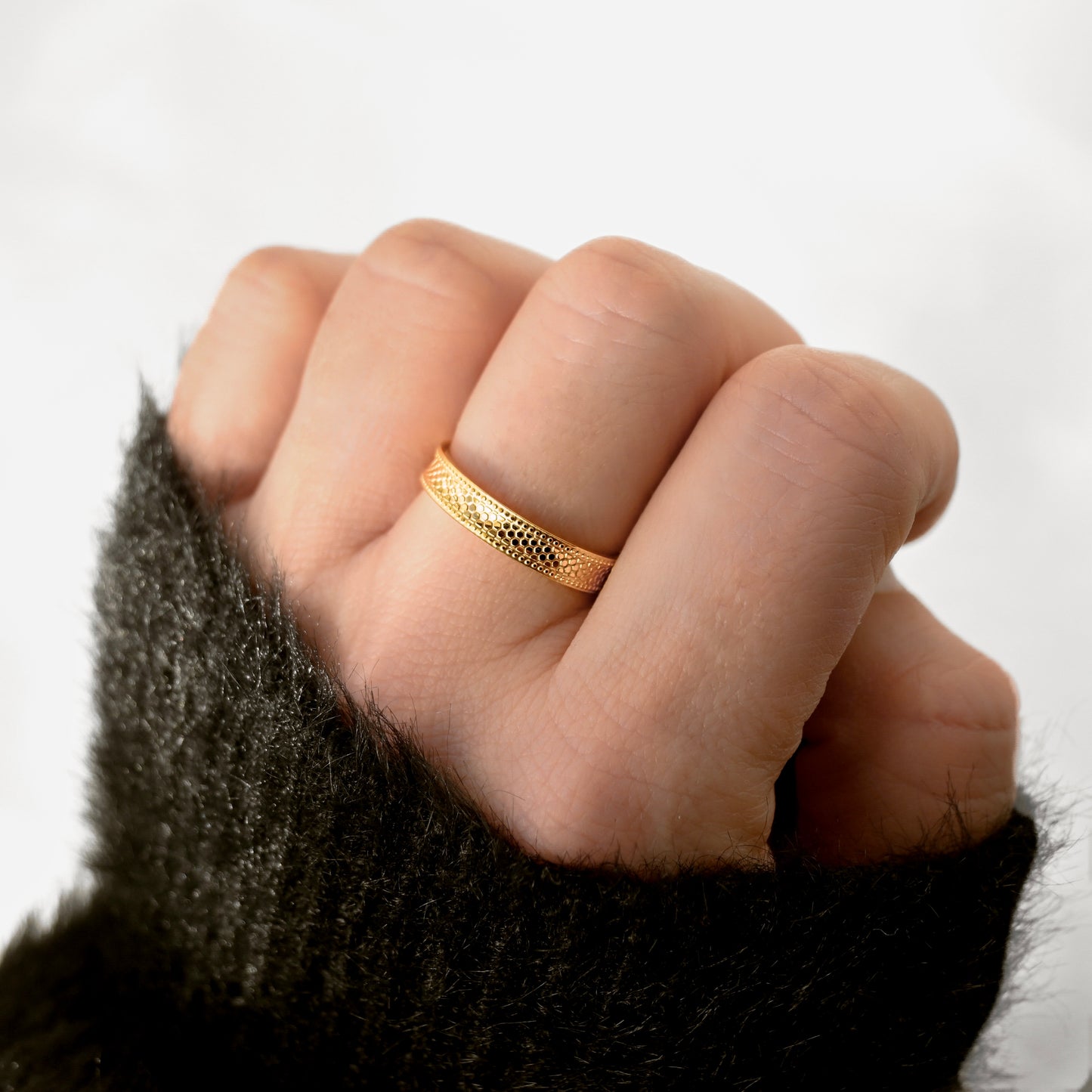 A close-up of a hand wearing a textured gold band ring, shown against a soft white background with a dark fuzzy sleeve visible at the wrist.