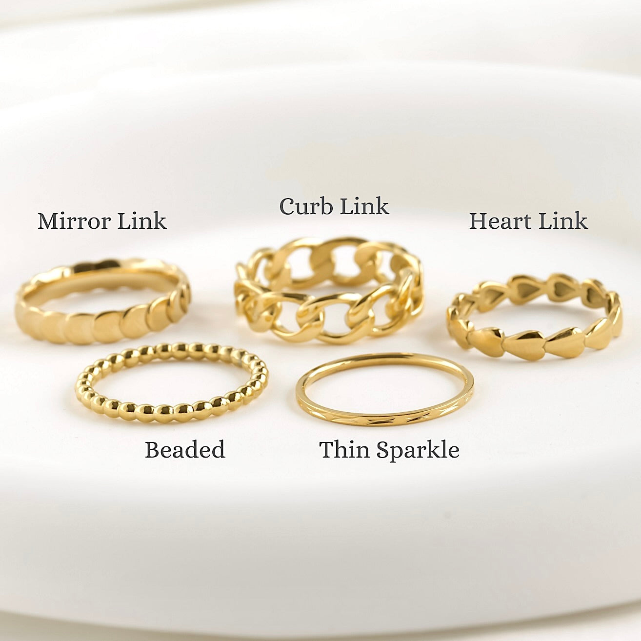 Set of gold rings with different designs on a white background including, scalloped, bold curb chain, heart chain, beaded and thin sparkle designs