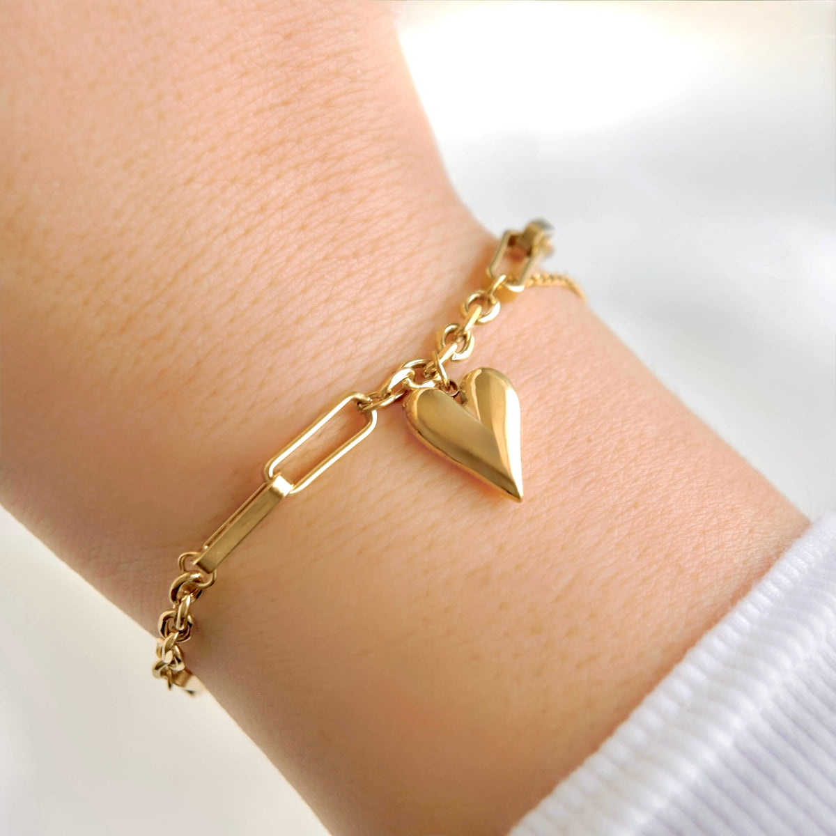 A gold chain bracelet with a polished heart charm and a paperclip link detail, shown worn on a person's wrist.