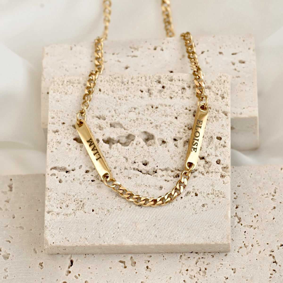 A dainty gold cuban chain necklace with two vertical bar charms engraved with 'LIAM' and 'ELOISE', displayed on a textured beige stone block.
