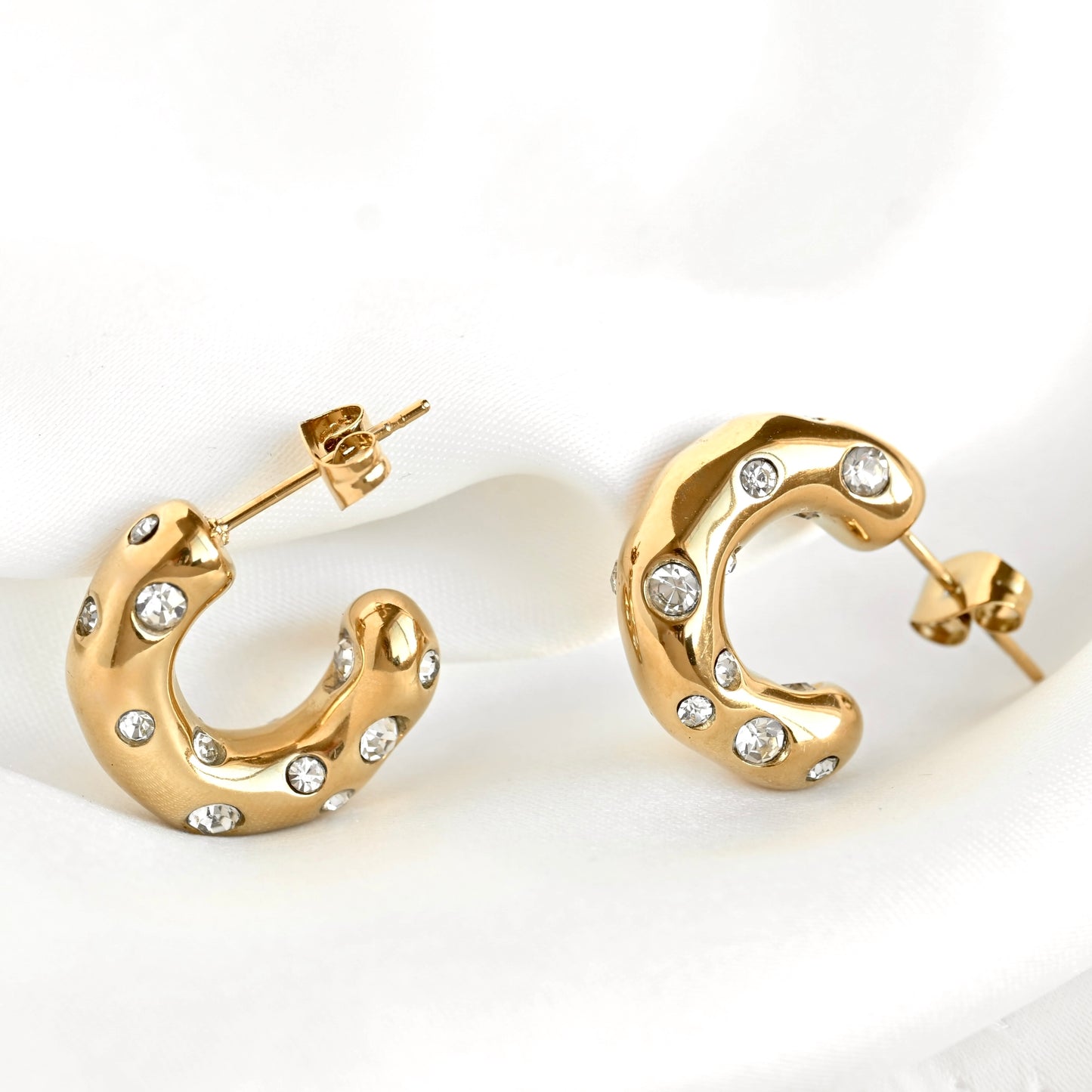 A pair of chunky gold crescent-shaped hoop earrings decorated with small clear stones, displayed on a soft white fabric background.
