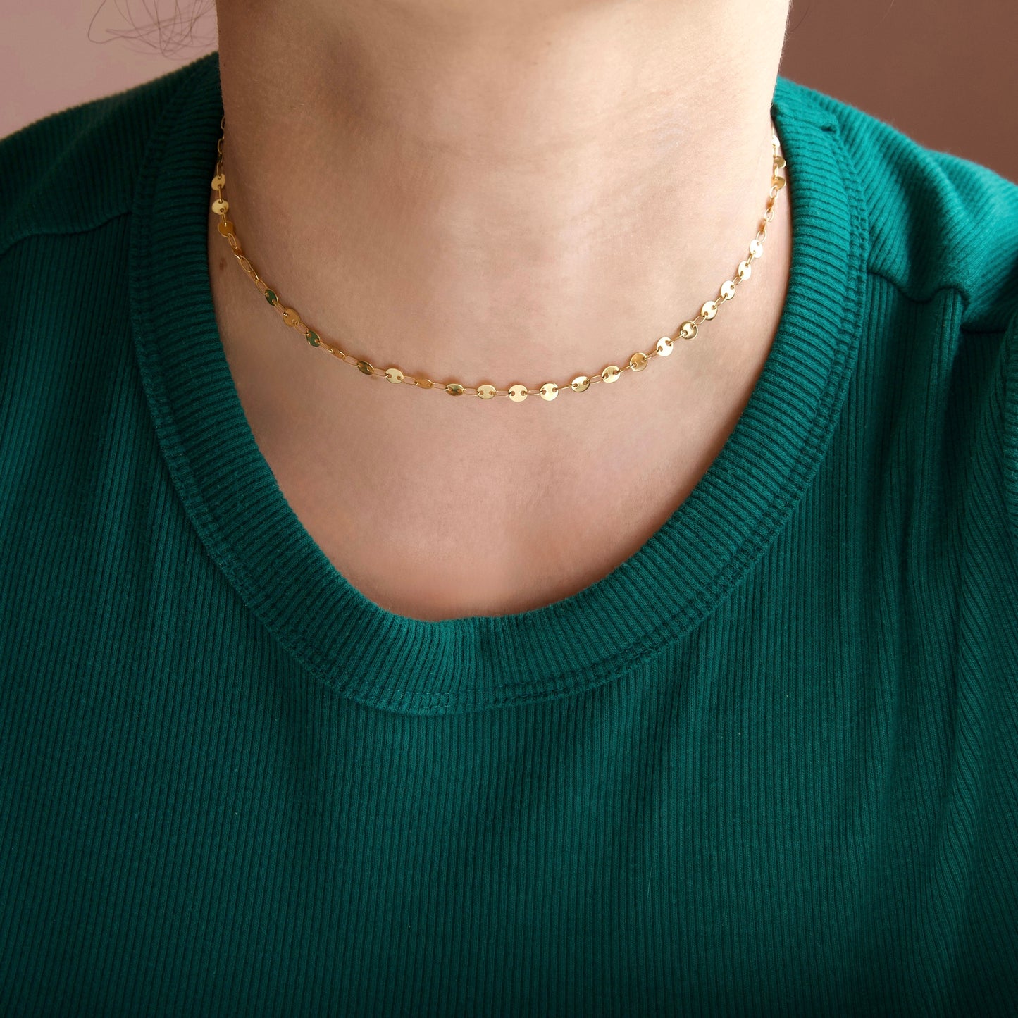 A close-up of a person wearing a delicate gold chain necklace with small faceted oval links, styled over a dark green ribbed crewneck top.
