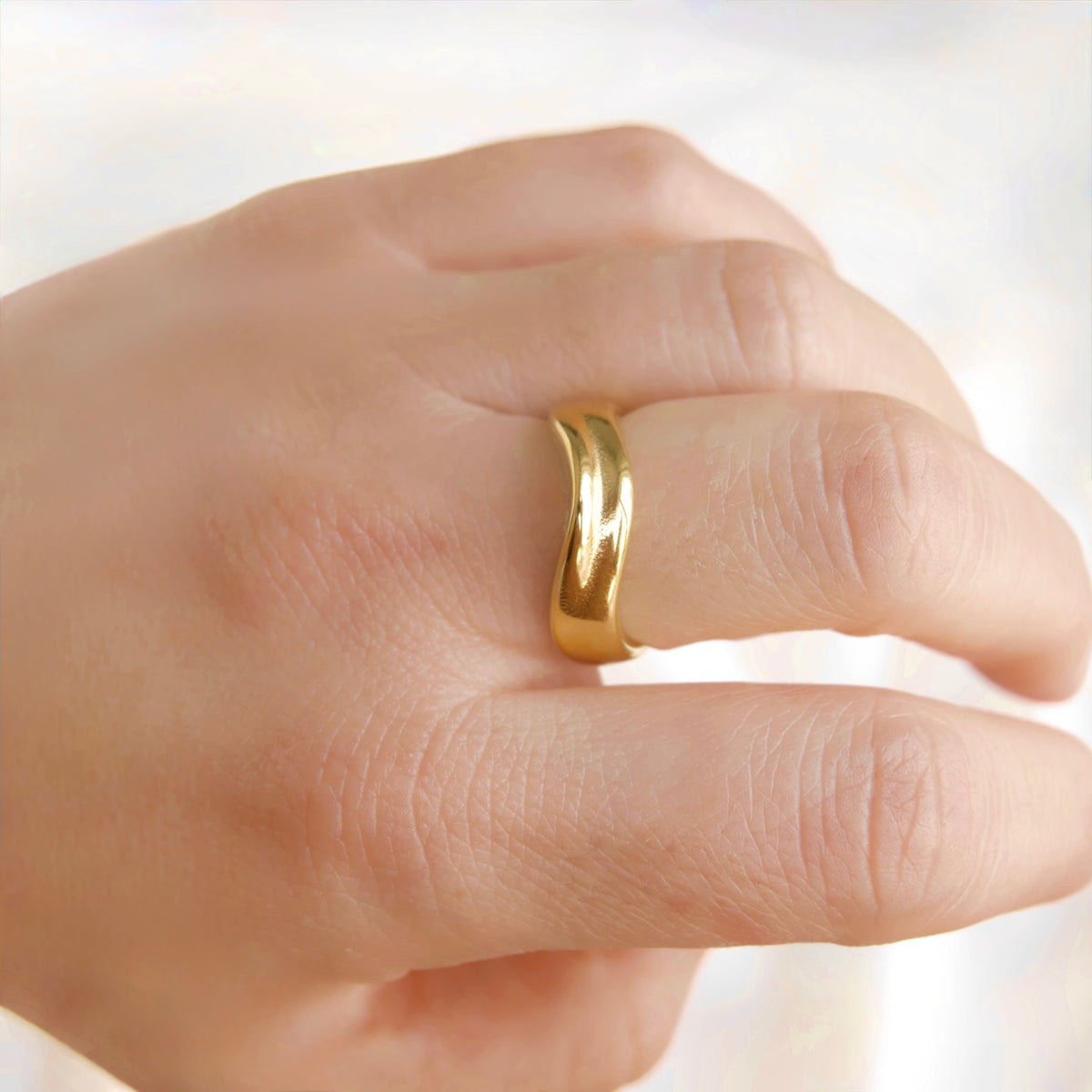 A close-up of a hand wearing a polished gold band ring with a wavy, undulating design.