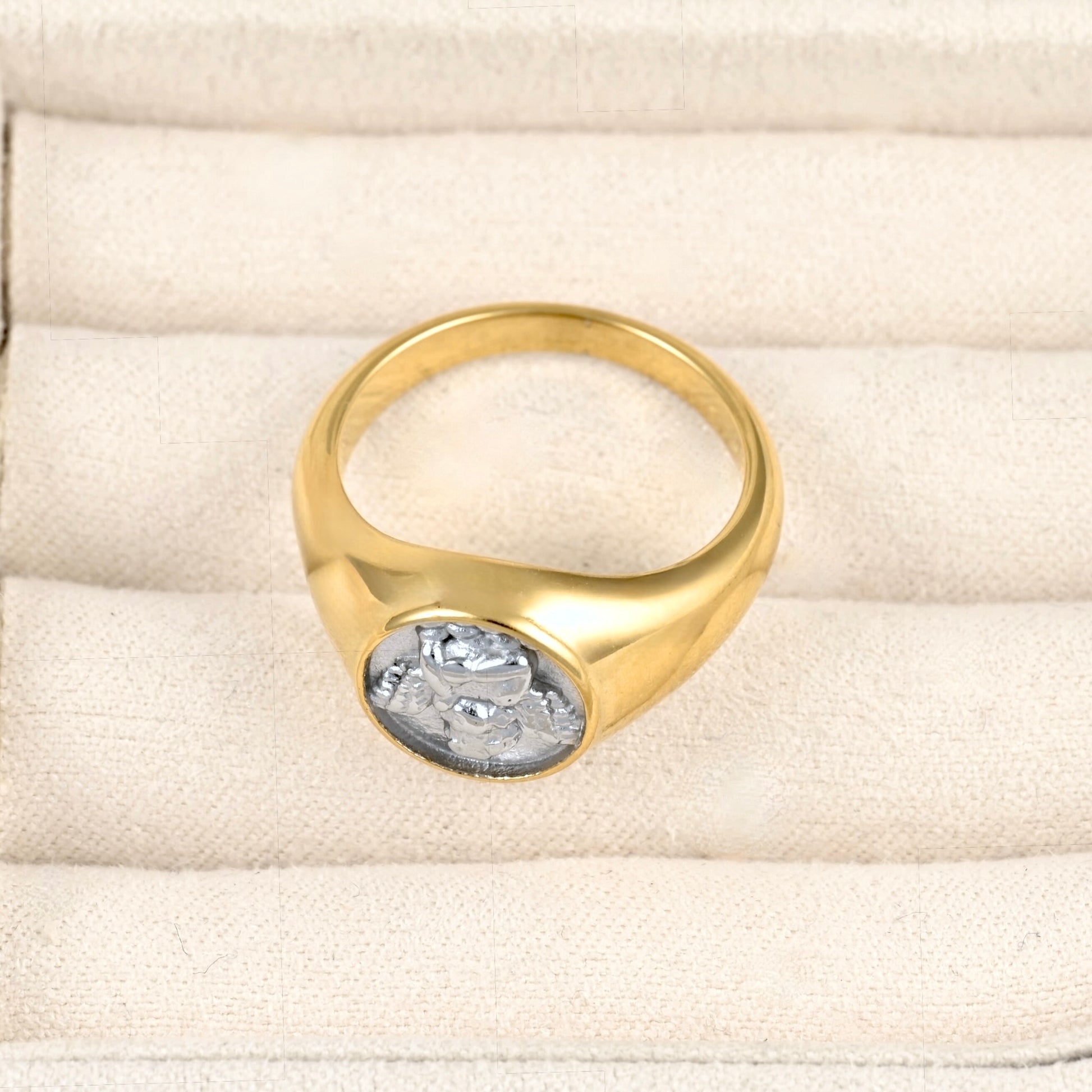 A gold signet ring with a silver-toned circular medallion featuring a profile bust, displayed on a cream-colored textured ring box.