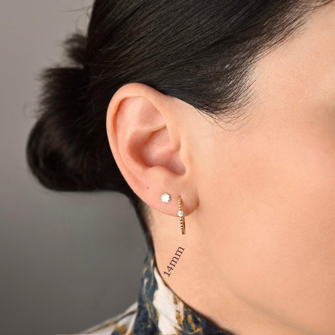 Close-up of a person's ear wearing medium gold beaded huggie hoop earrings with a tiny clear stone in the front. The text '14mm' is printed near the earlobe