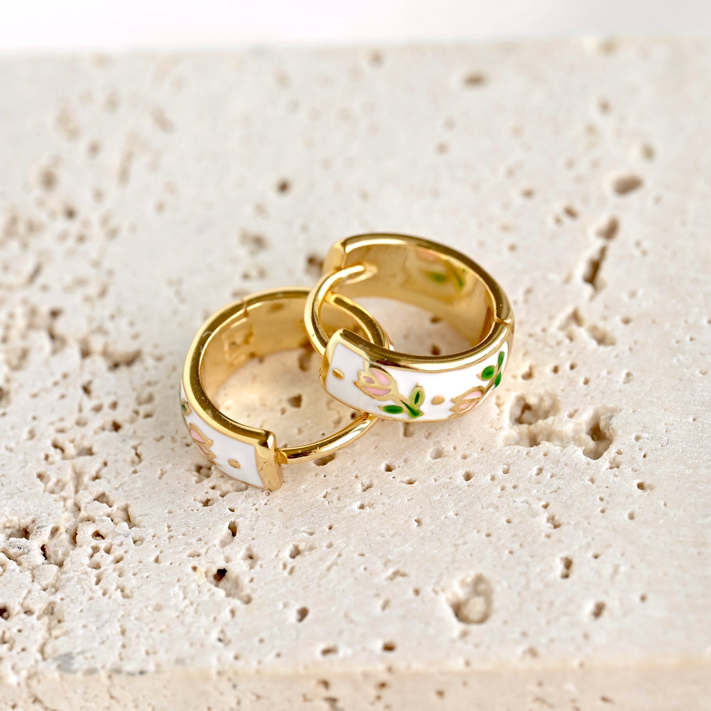 Two gold huggie hoop earrings with white enamel and a delicate floral pattern, resting on a textured beige stone surface.