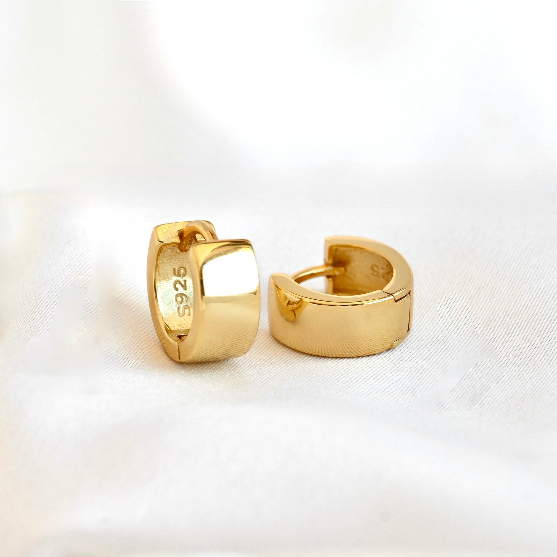 A pair of polished yellow gold huggie hoop earrings with '925' sterling silver purity stamped on the inside. They are displayed on a soft, white textured fabric background.