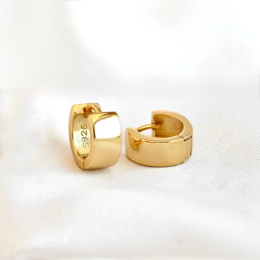 A pair of polished yellow gold huggie hoop earrings with '925' sterling silver purity stamped on the inside. They are displayed on a soft, white textured fabric background.
