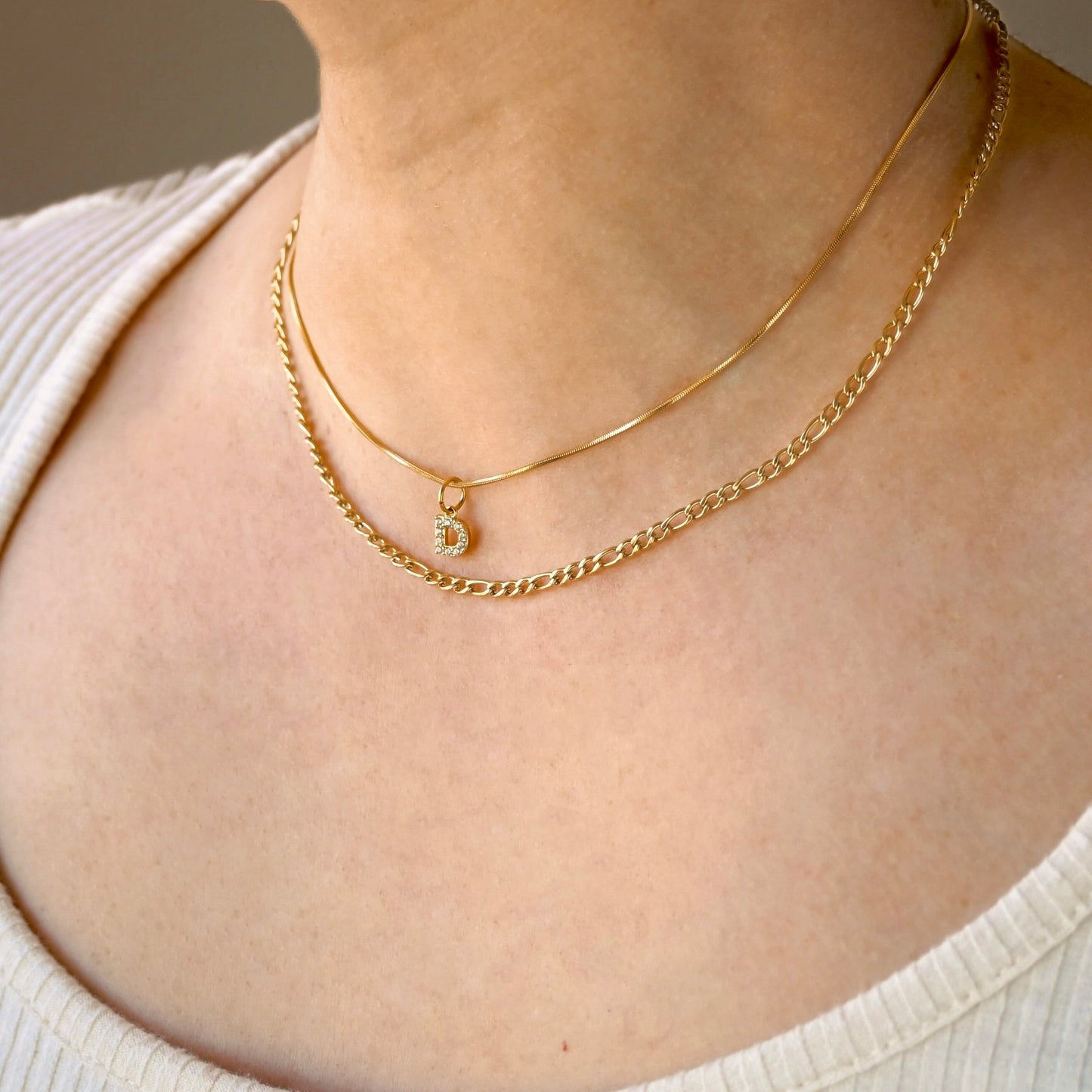 Gold figaro necklace layered with a skinny snake chain with a diamond letter charm on a person wearing a white sweater.