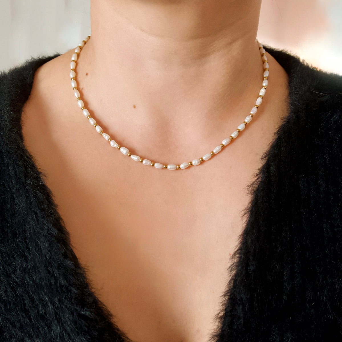 A close-up of a person wearing a delicate gold chain necklace with irregular white pearls, styled with a black fuzzy knit top.