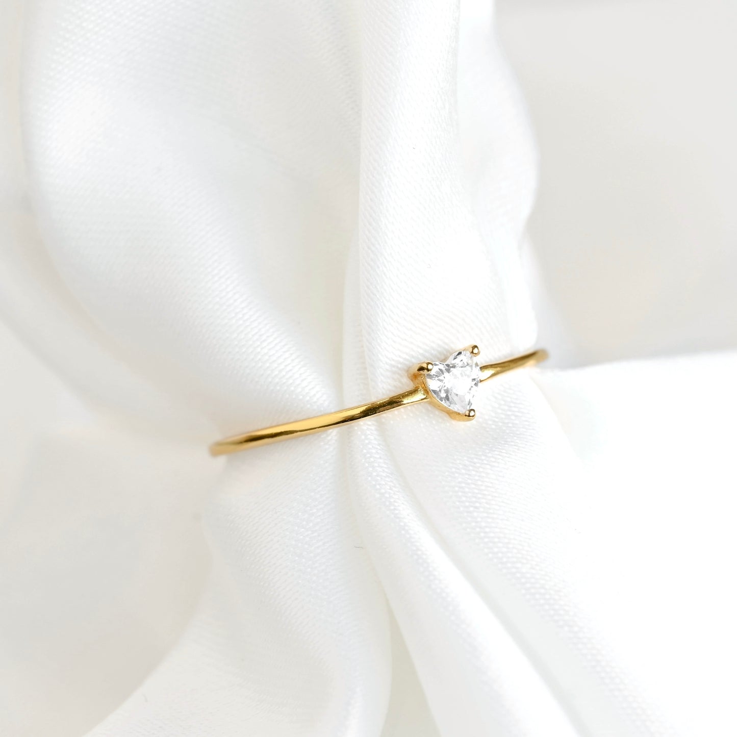 A delicate gold ring with a small heart-cut stone, displayed on soft white draped fabric.
