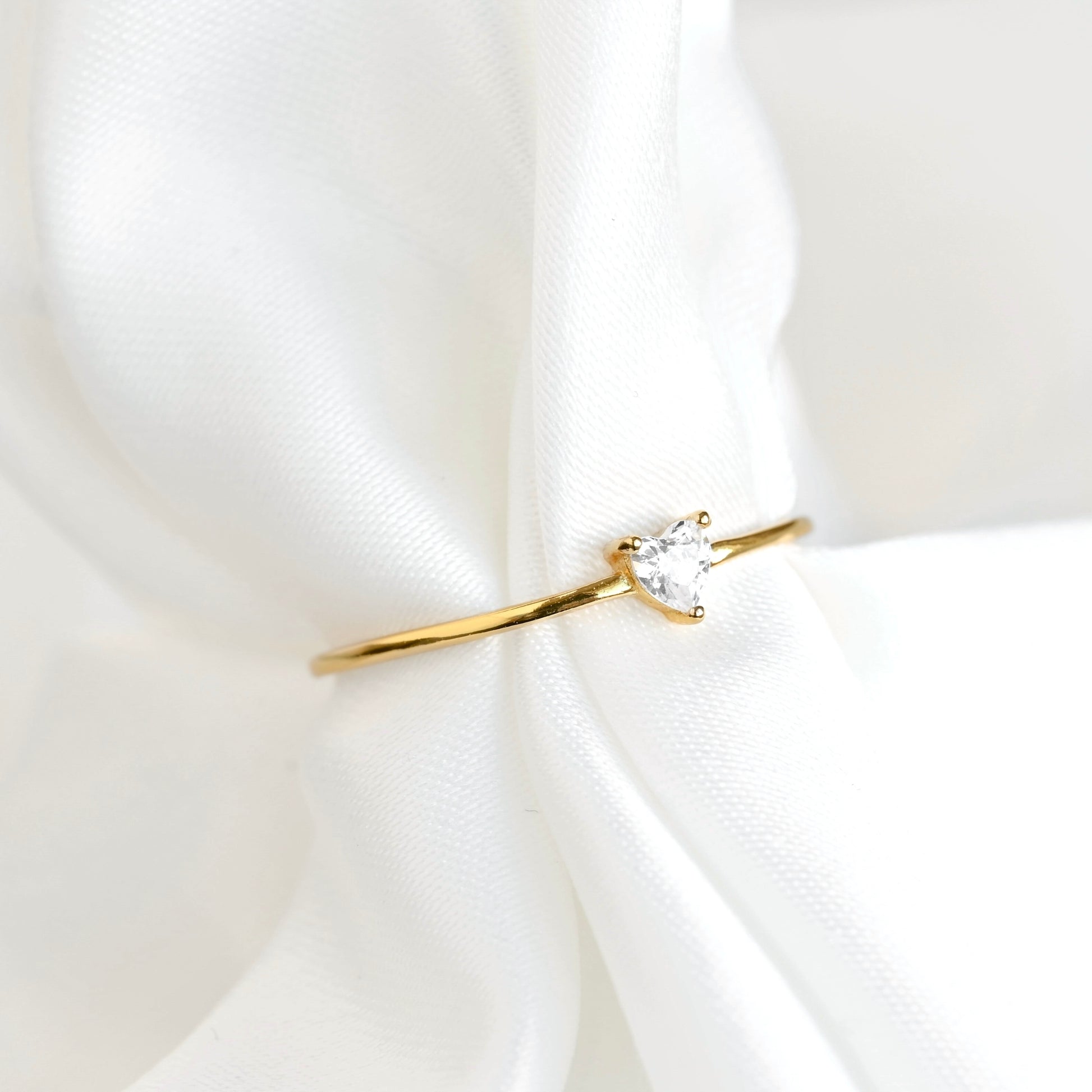 A delicate gold ring with a small heart-cut stone, displayed on soft white draped fabric.