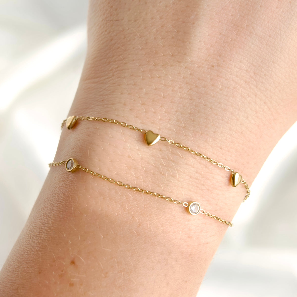 Double gold chain bracelets with small heart and round bezel details, shown worn on a person's wrist against a soft white background.