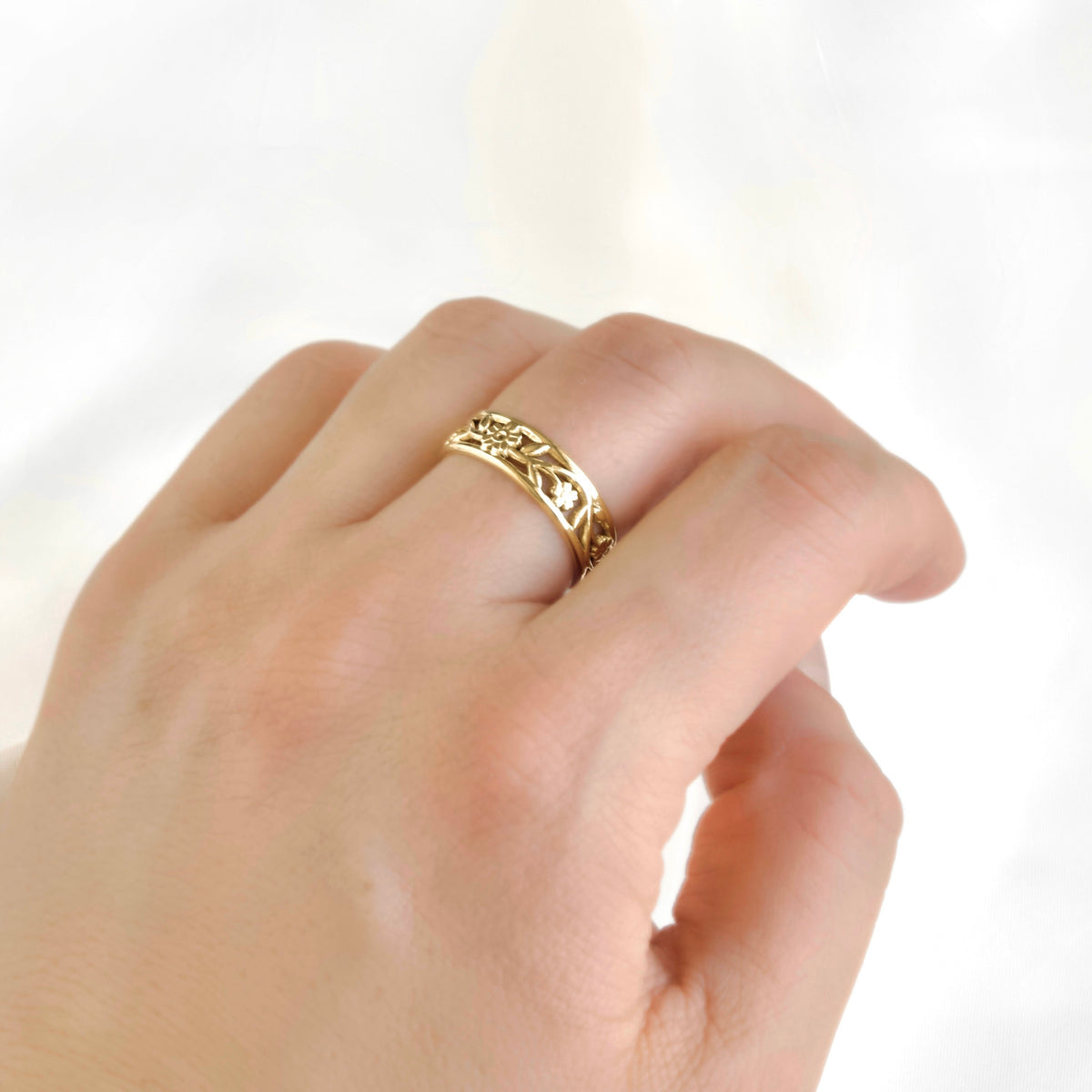 A dainty gold band with cut-out filigree pattern with flowers and leafs on a women's middle finger.