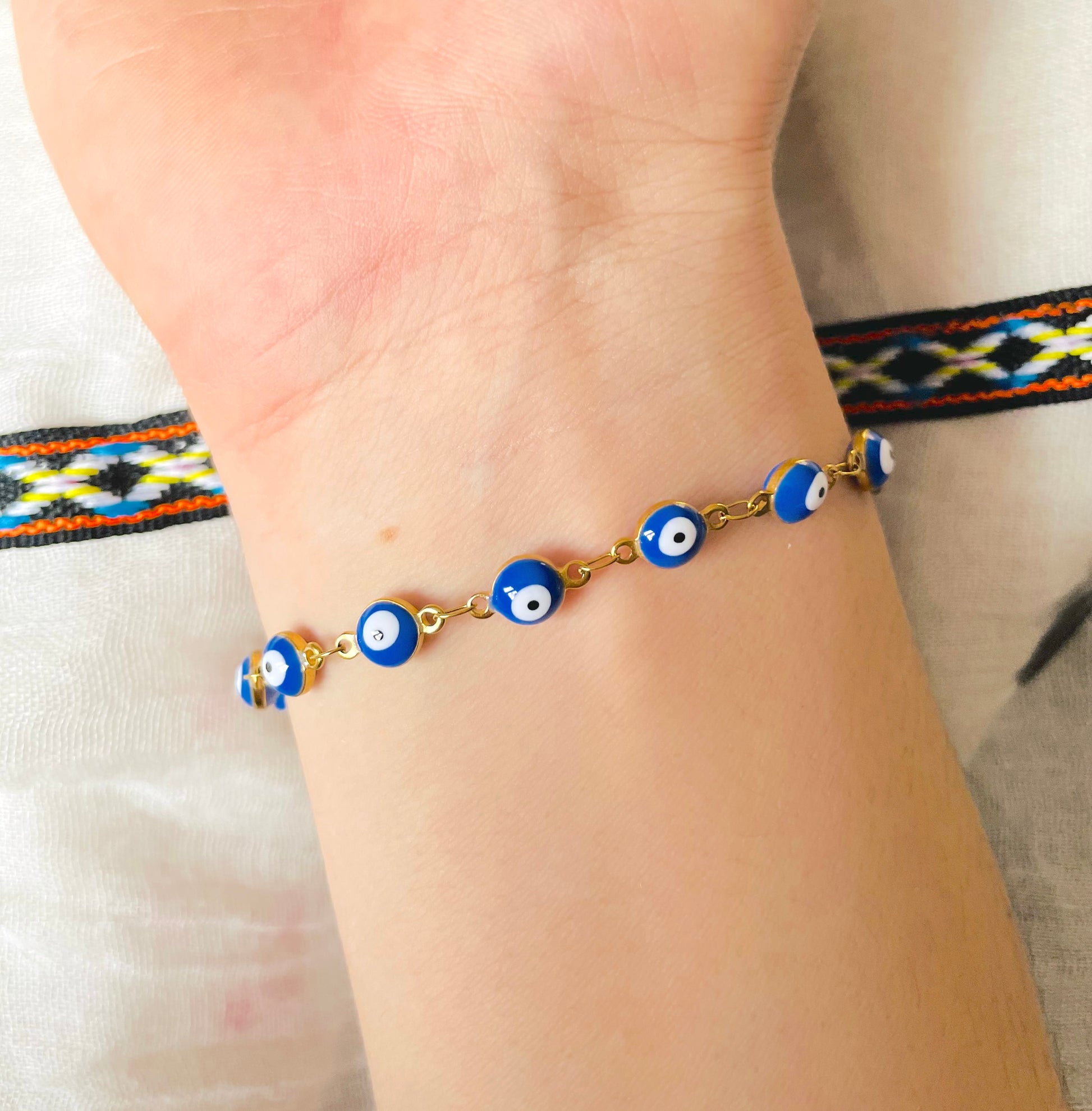 Gold bracelet with blue evil eye beads on a wrist, with a colorful patterned band in the background.