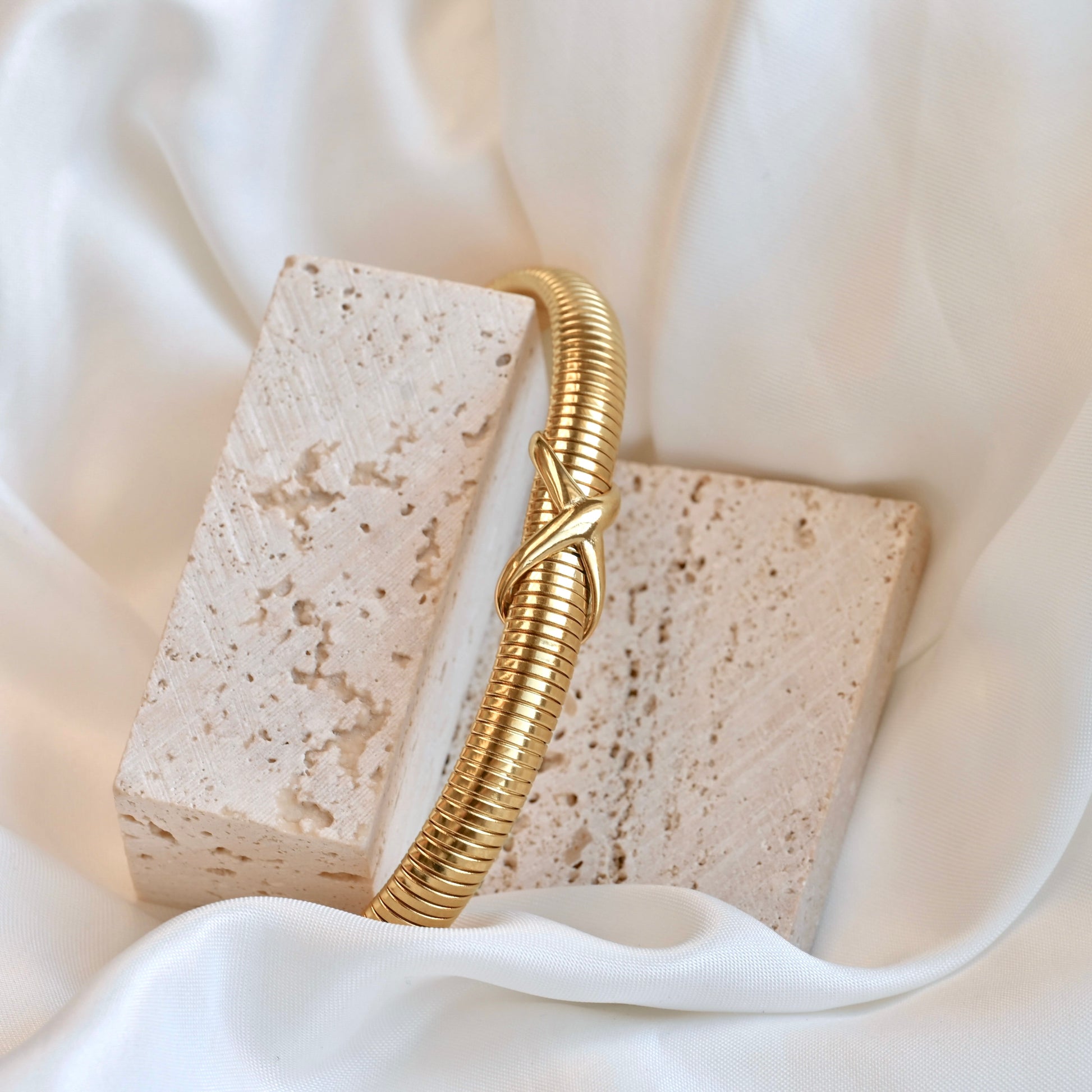 Gold knotted bangle bracelet on a textured stone block with a soft white fabric background