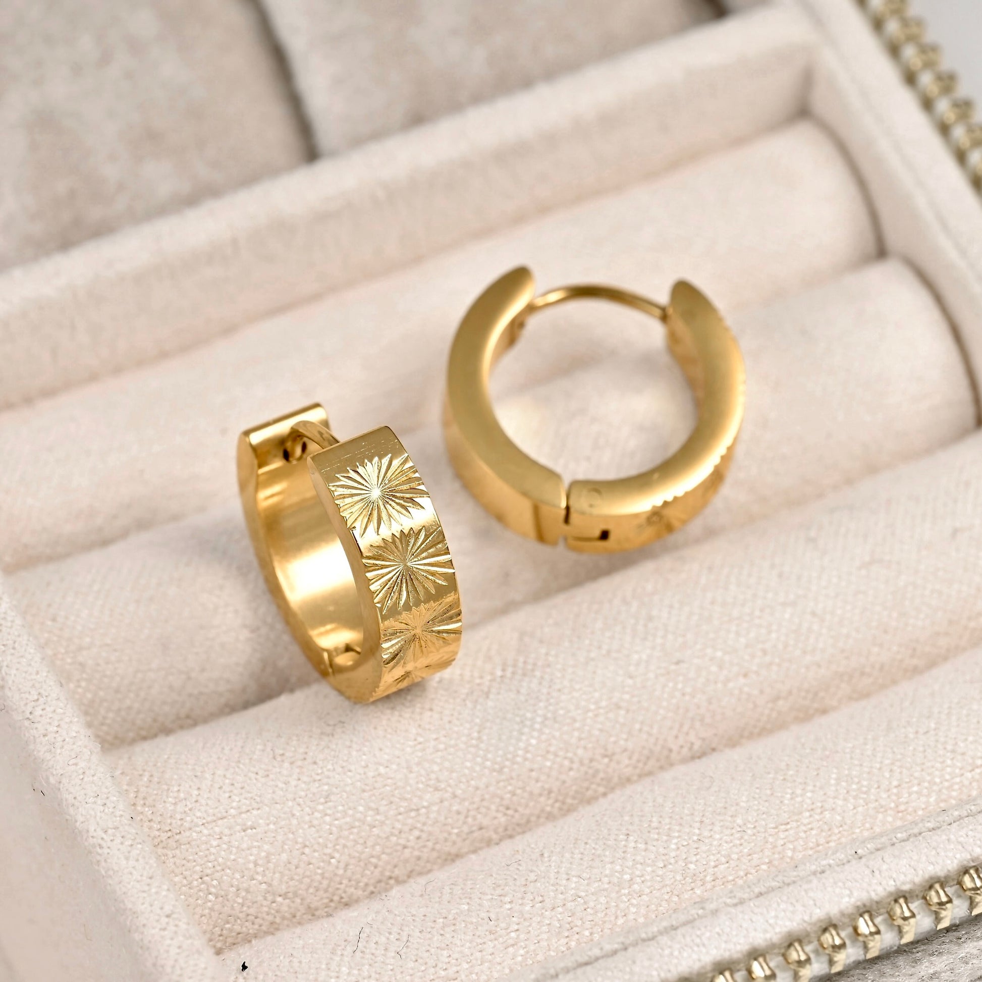 A pair of gold huggie hoop earrings with a sunburst star pattern, one upright, one resting on a soft beige velvet-lined jewelry box.
