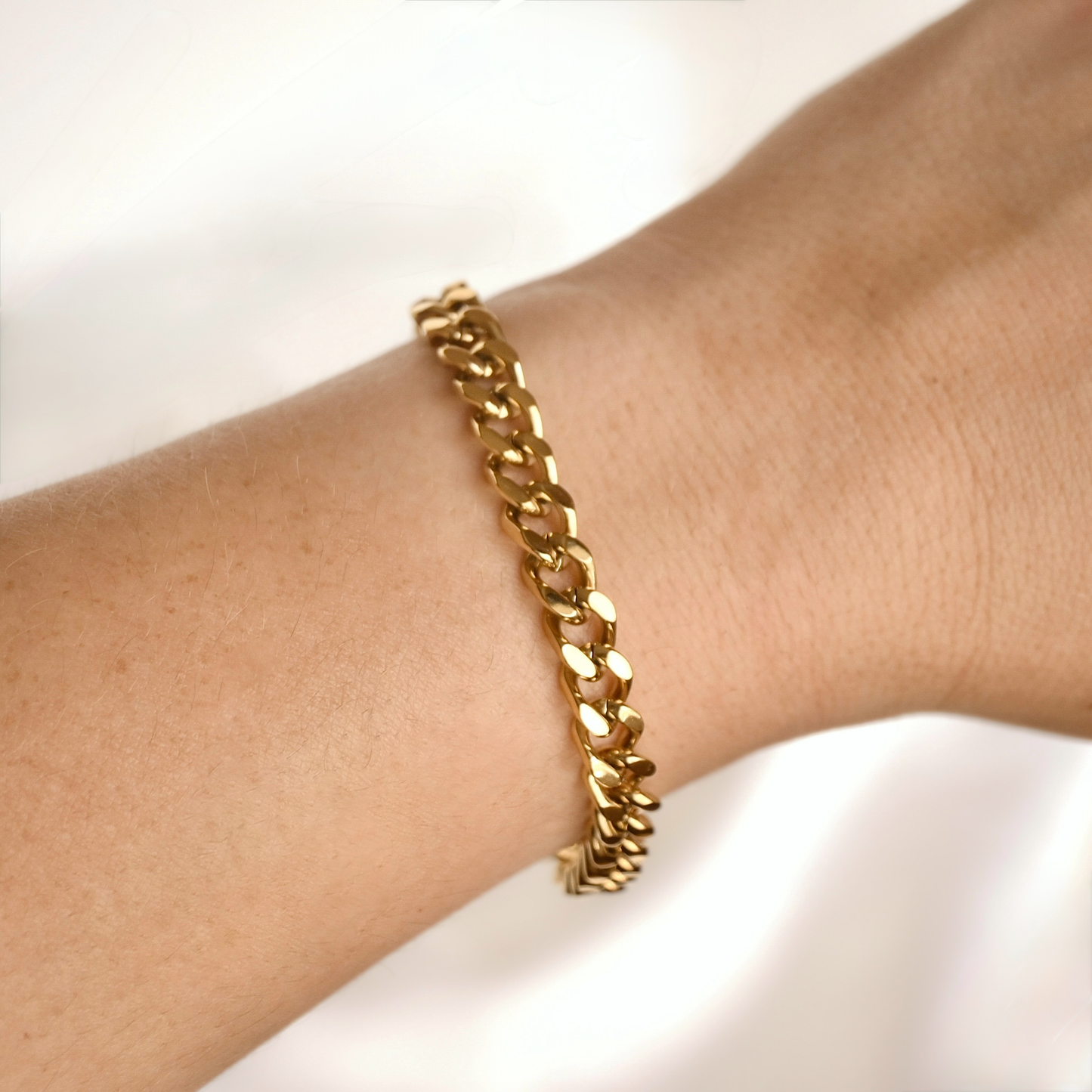 A close-up of a gold Cuban link chain bracelet worn on a person's wrist against a soft white background.