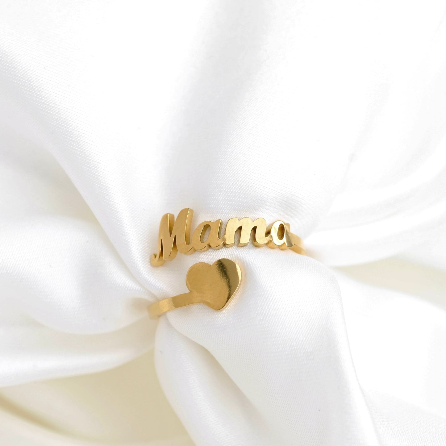 A gold-plated name ring spelling 'Mama' with a small heart charm, displayed on soft white satin fabric.