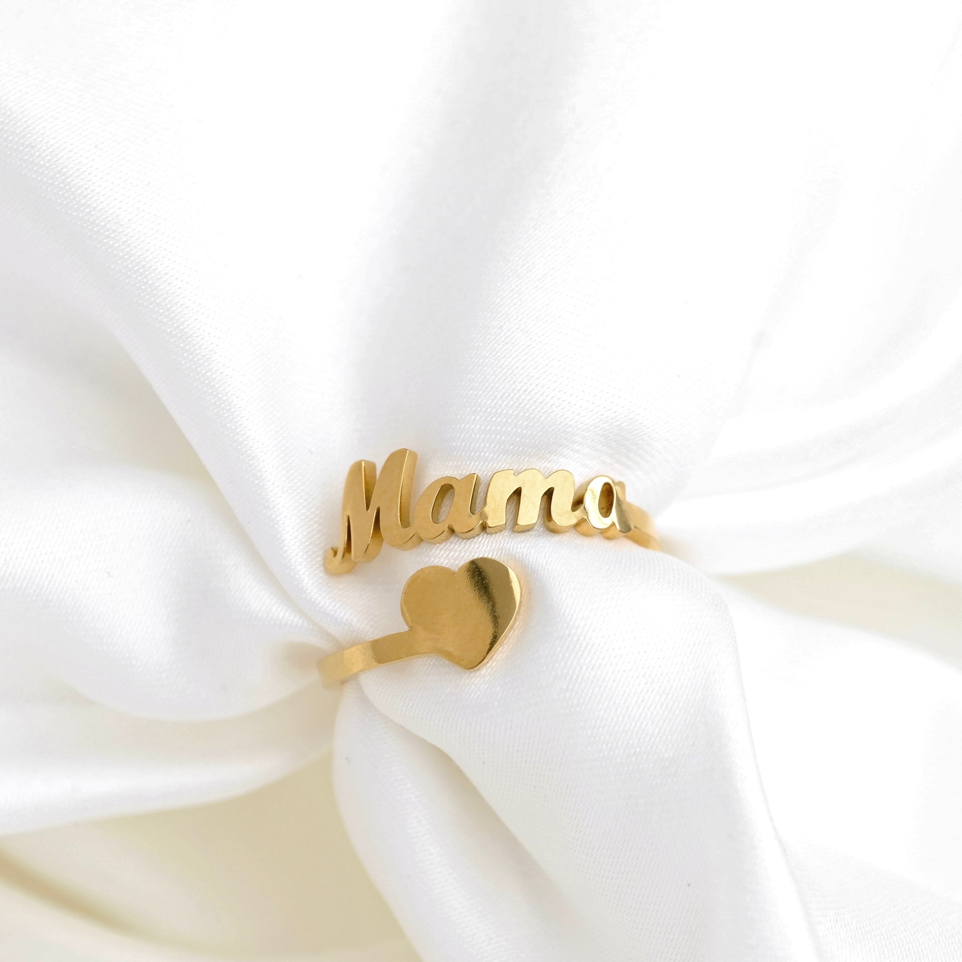 A gold-plated name ring spelling 'Mama' with a small heart charm, displayed on soft white satin fabric.
