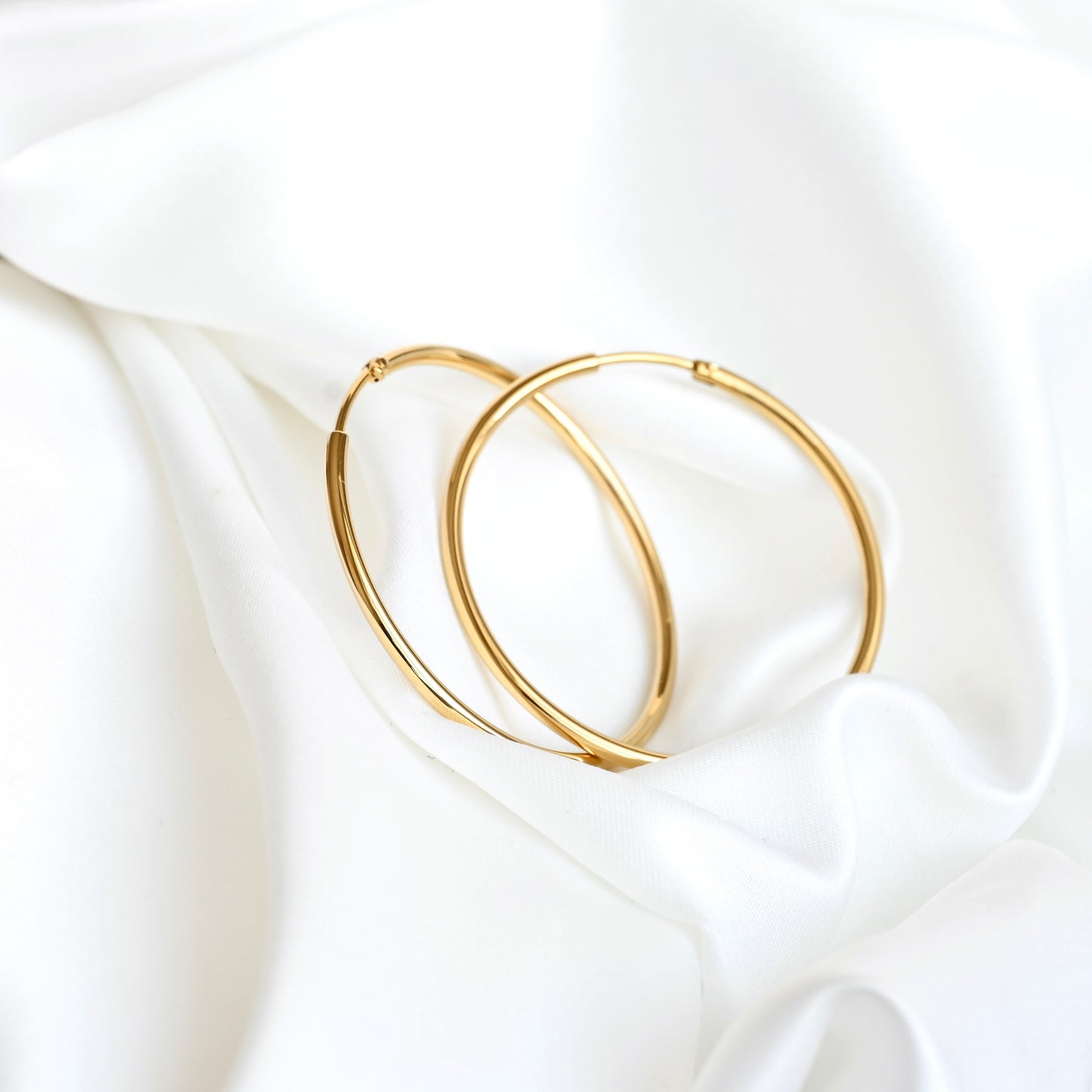 Two thin gold hoop earrings resting on draped white satin fabric.