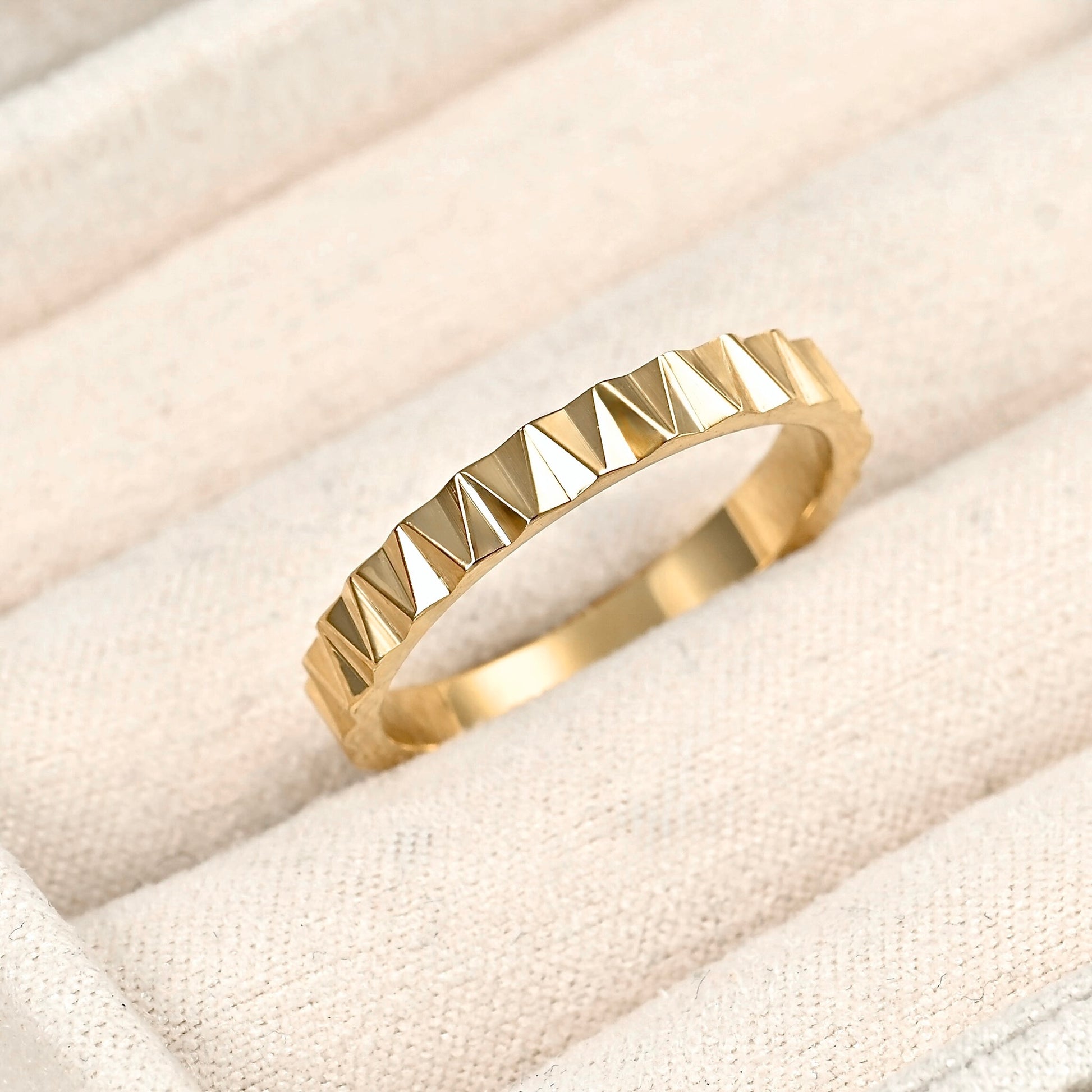 A gold ring with a textured, faceted triangular pattern, resting on a beige fabric jewelry display.
