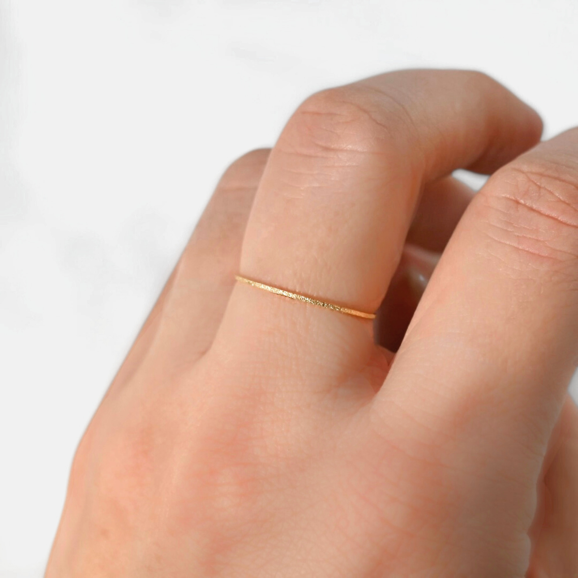A close-up of a hand wearing a delicate, thin gold ring on one finger against a soft, light gray background.