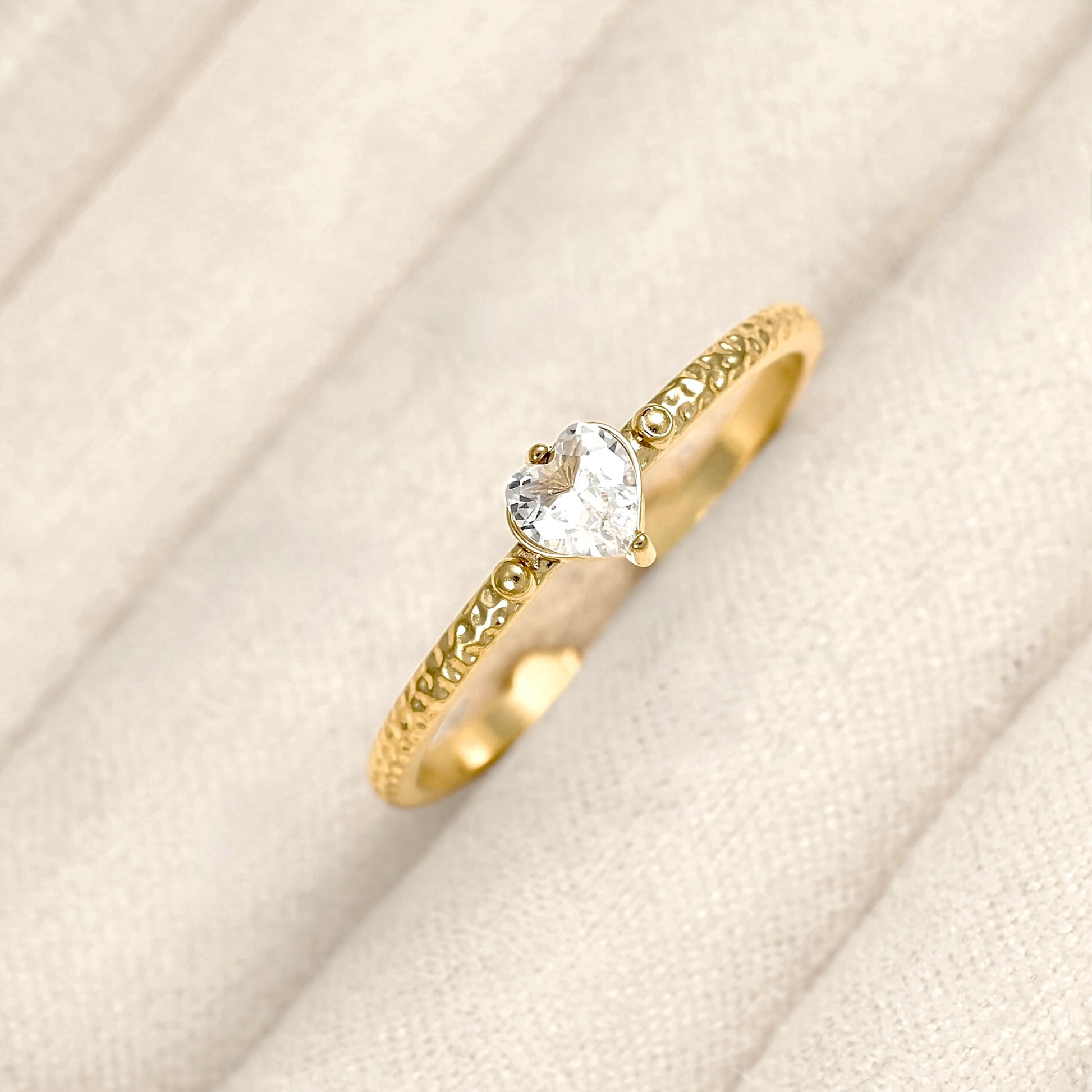 A gold ring with an engraved band and a clear heart-shaped gemstone, displayed on a textured cream fabric background.