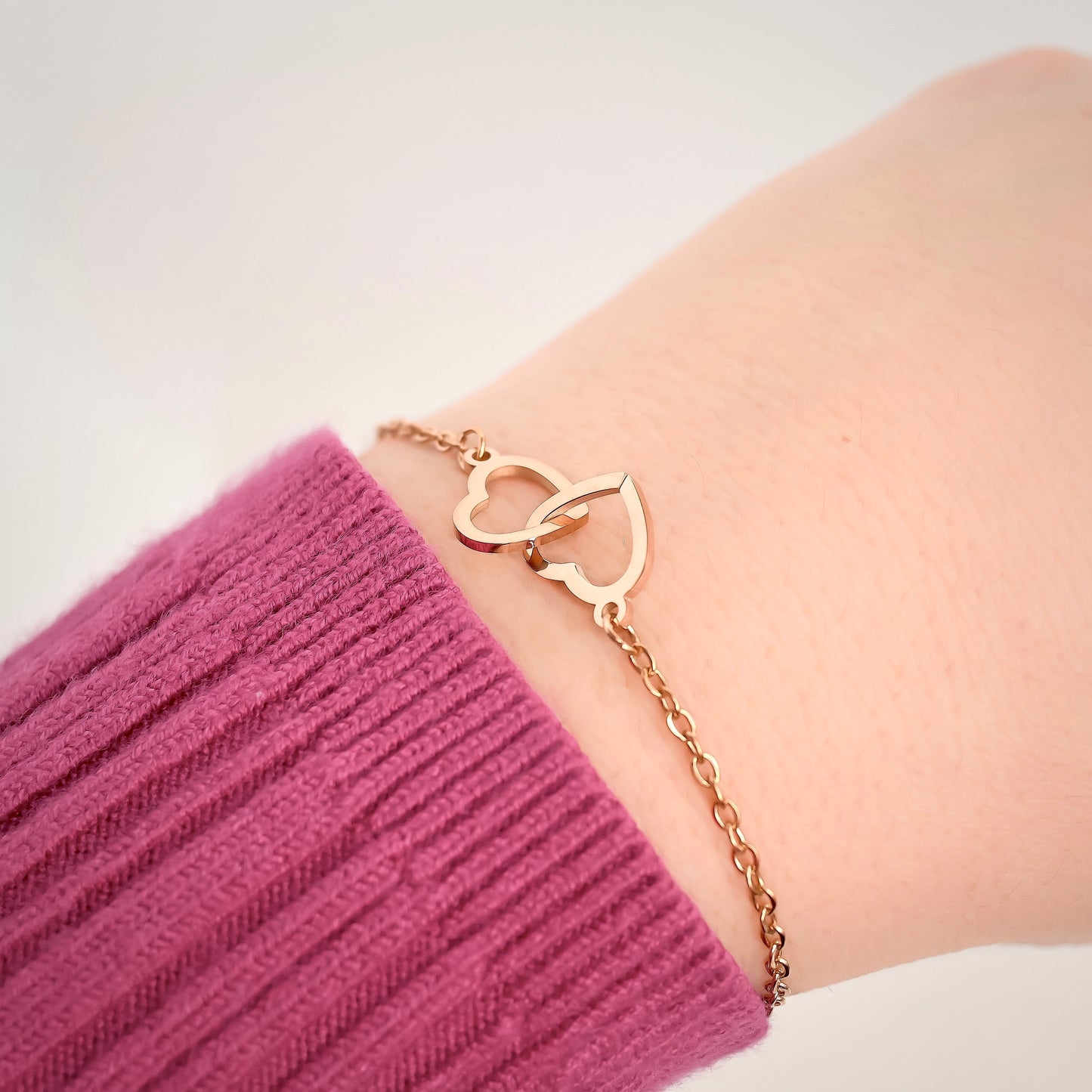 Rose gold interlocking heart bracelet on a wrist with a pink sleeve against a white background