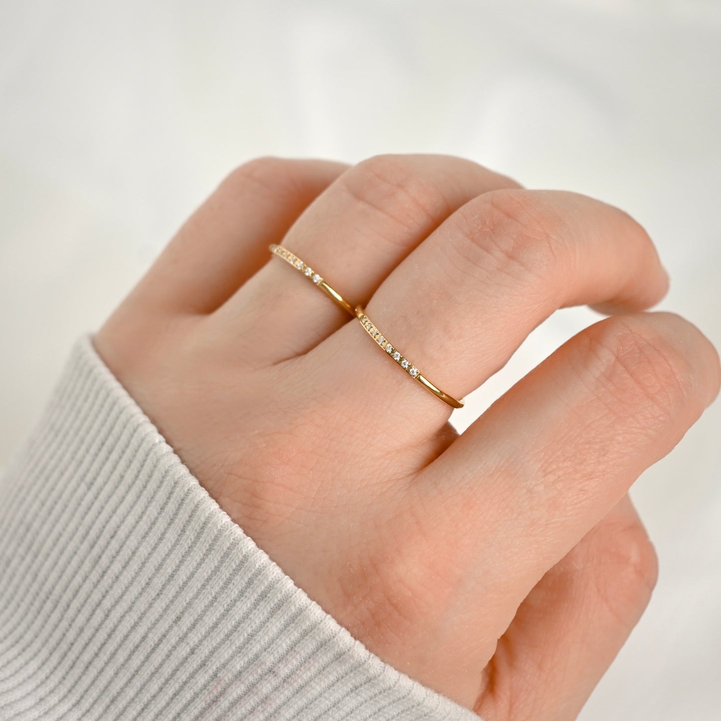 A close-up of a hand wearing two delicate gold rings,which feature small clear stones, set against a soft, light gray background.