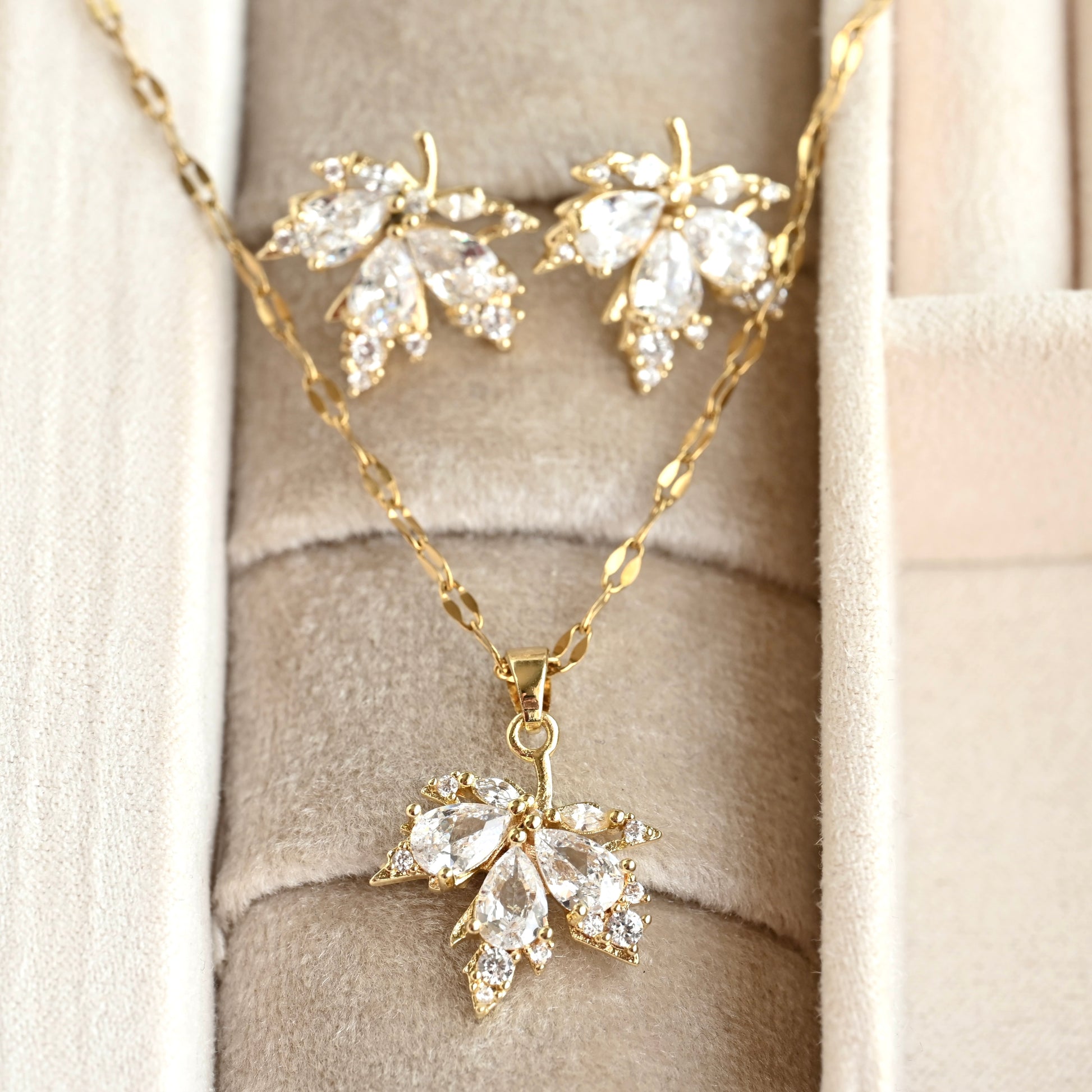 Gold-tone leaf-shaped pendant necklace and matching stud earrings with clear teardrop gemstones, displayed on a beige jewelry box.