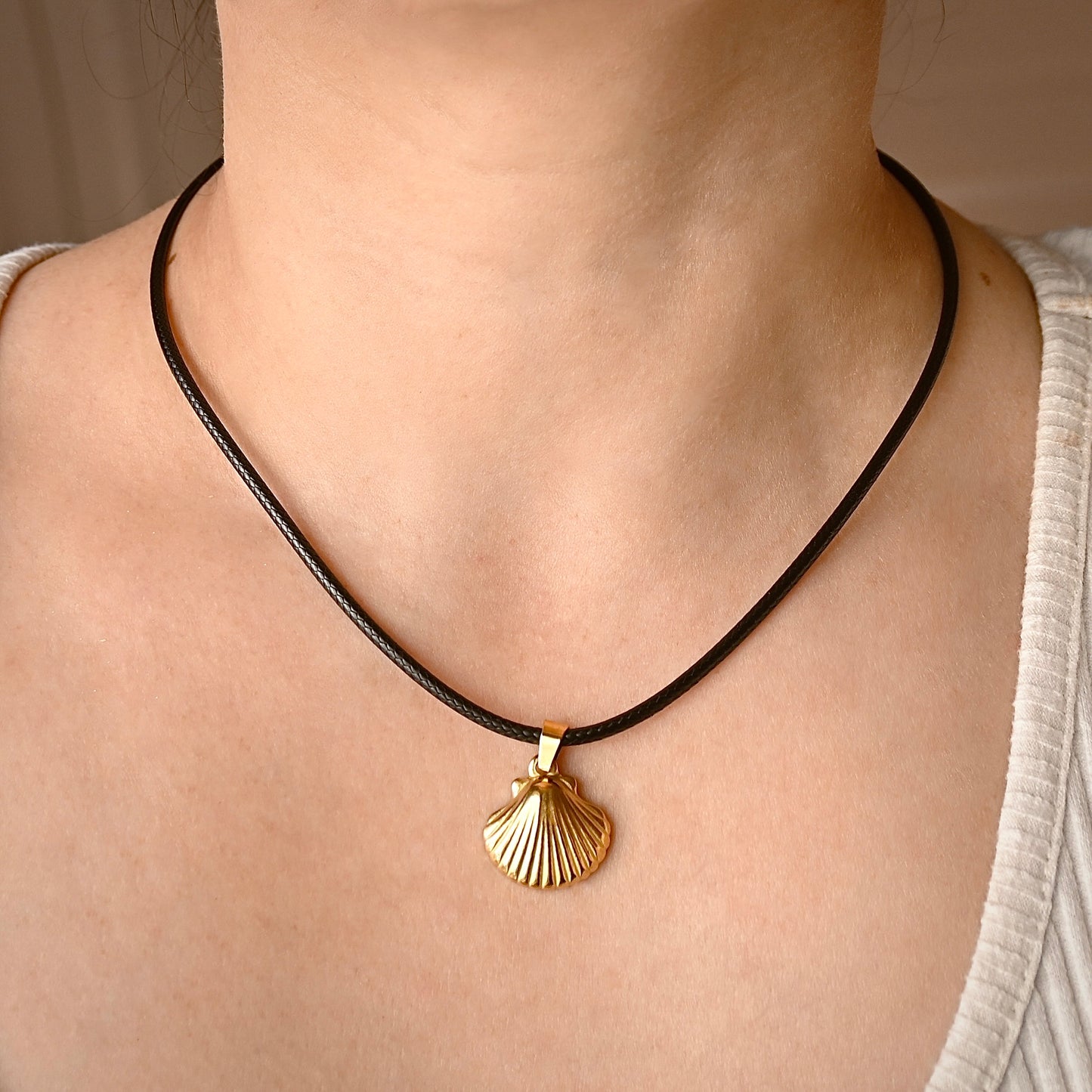 A close-up front view of a person's neck and chest wearing a black cord necklace with a gold shell-shaped pendant.