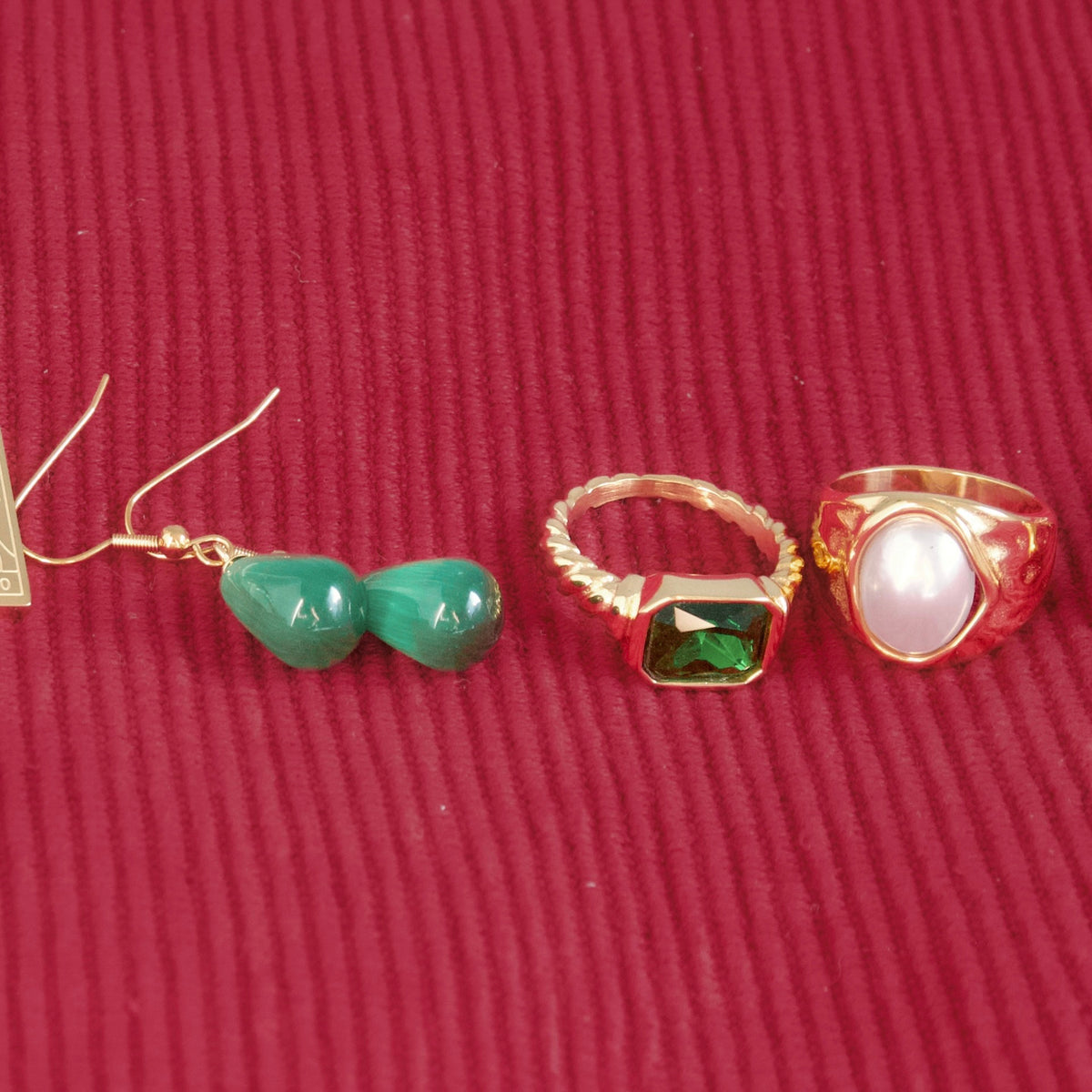 A jewelry set featuring a pair of gold dangle earrings with green beads, a gold ring with a green faceted gemstone, and a gold signet ring with a white oval stone, all arranged on a textured red background.