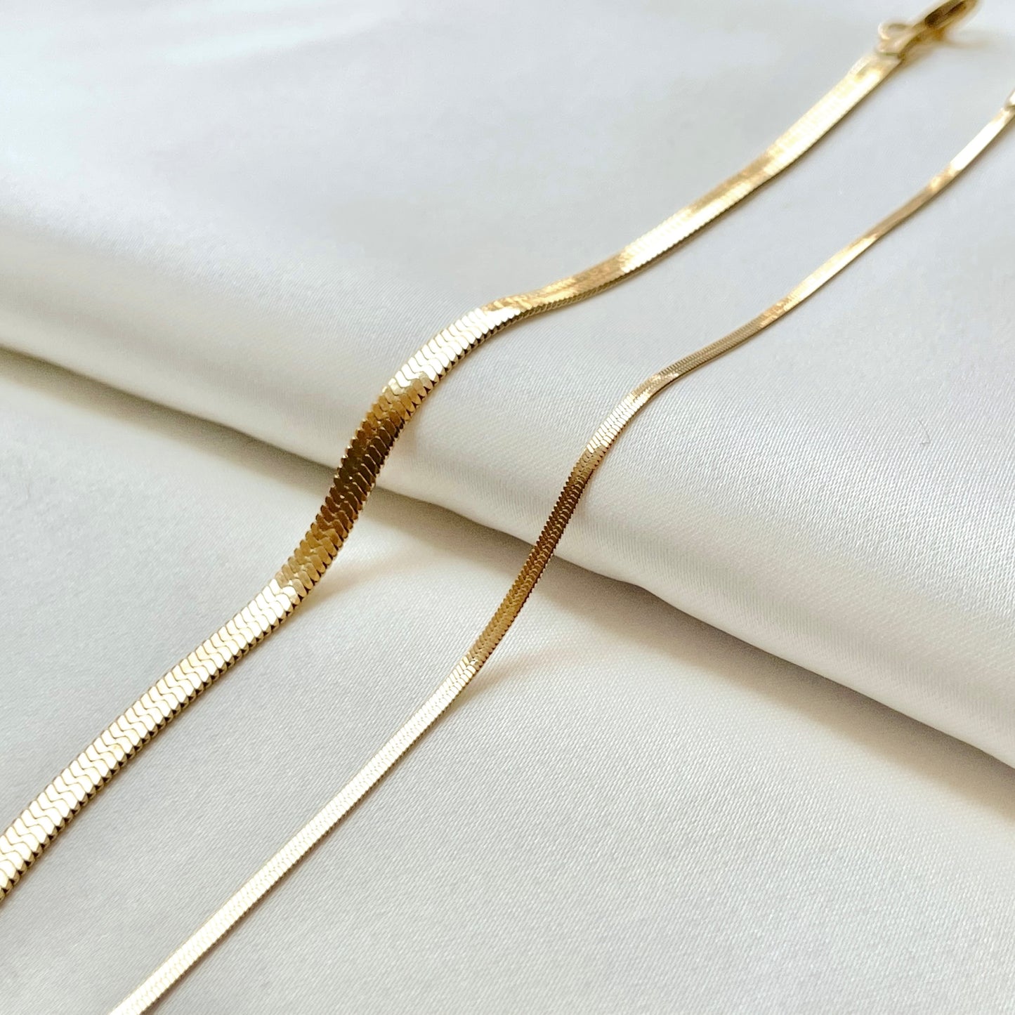 Flat gold snake chains, one 4mm one 2mm, on a white fabric background