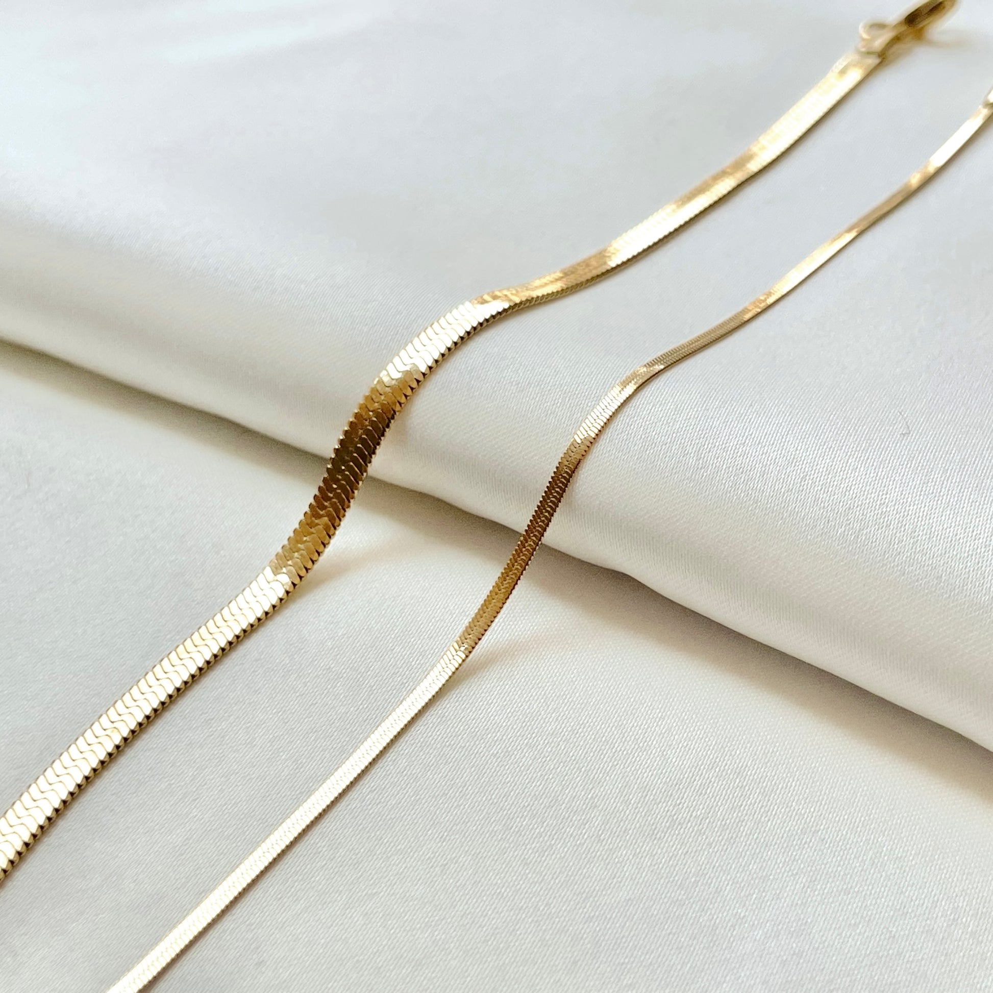Flat gold snake chains, one 4mm one 2mm, on a white fabric background