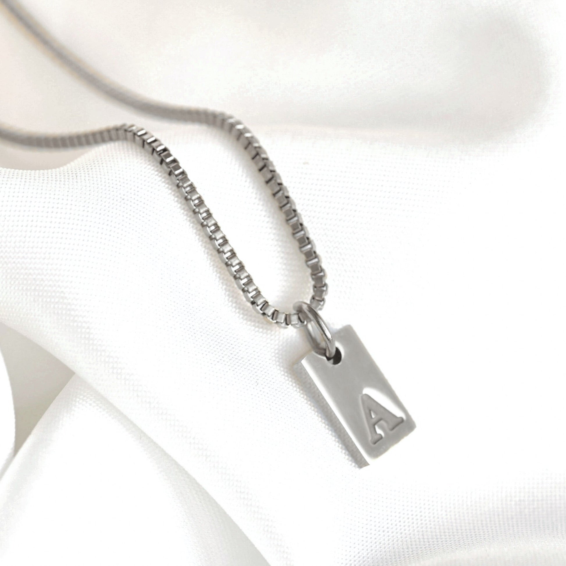 Silver box chain necklace with a rectengular initial pendant on a white satin background